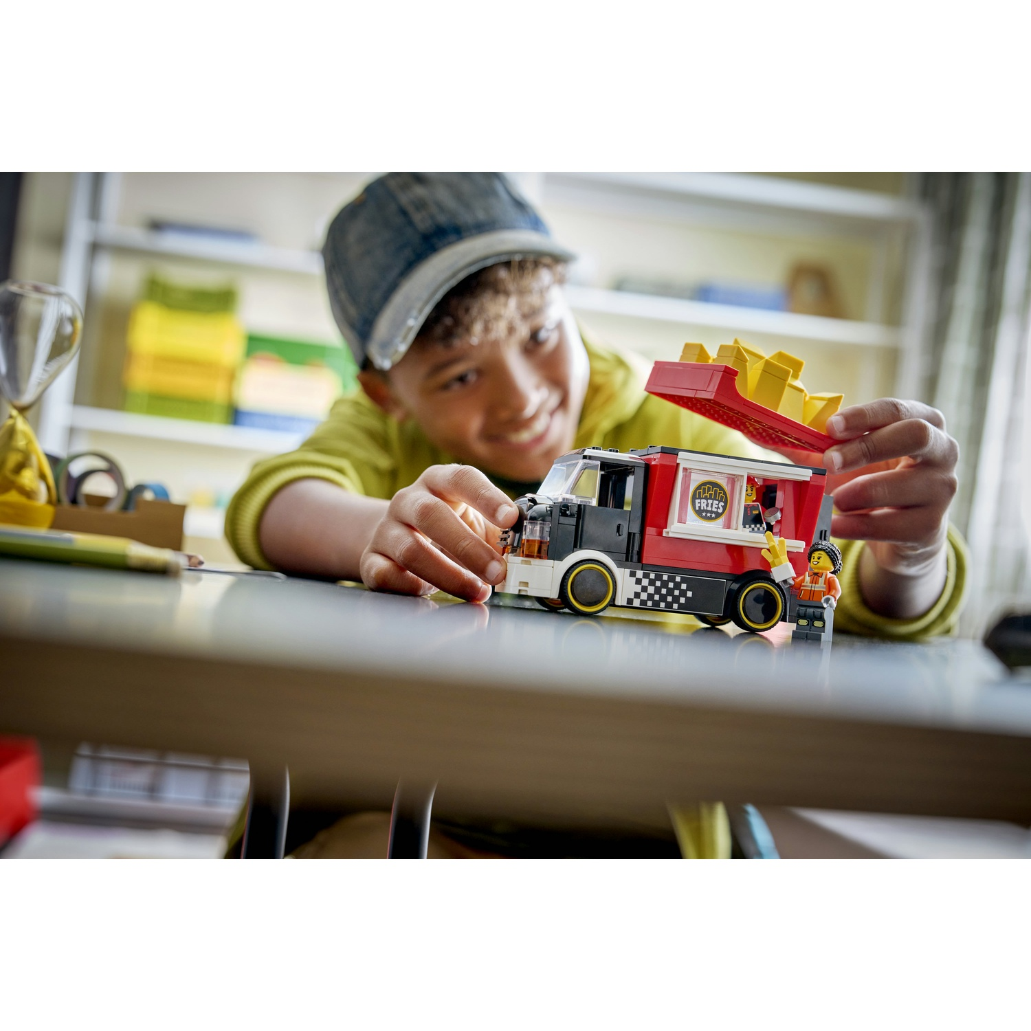 Un ragazzo sorride, gioca con un camioncino LEGO. Il camion è rosso e nero, con un contenitore di patatine fritte sopra. Una minifigure è accanto.