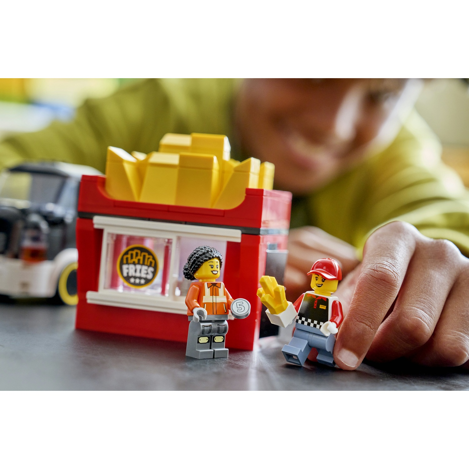 Un chiosco di patatine fritte Lego con due minifigure. Uno tiene una moneta, l'altro patatine fritte. Un'auto è visibile sullo sfondo, una mano di bambino è visibile.