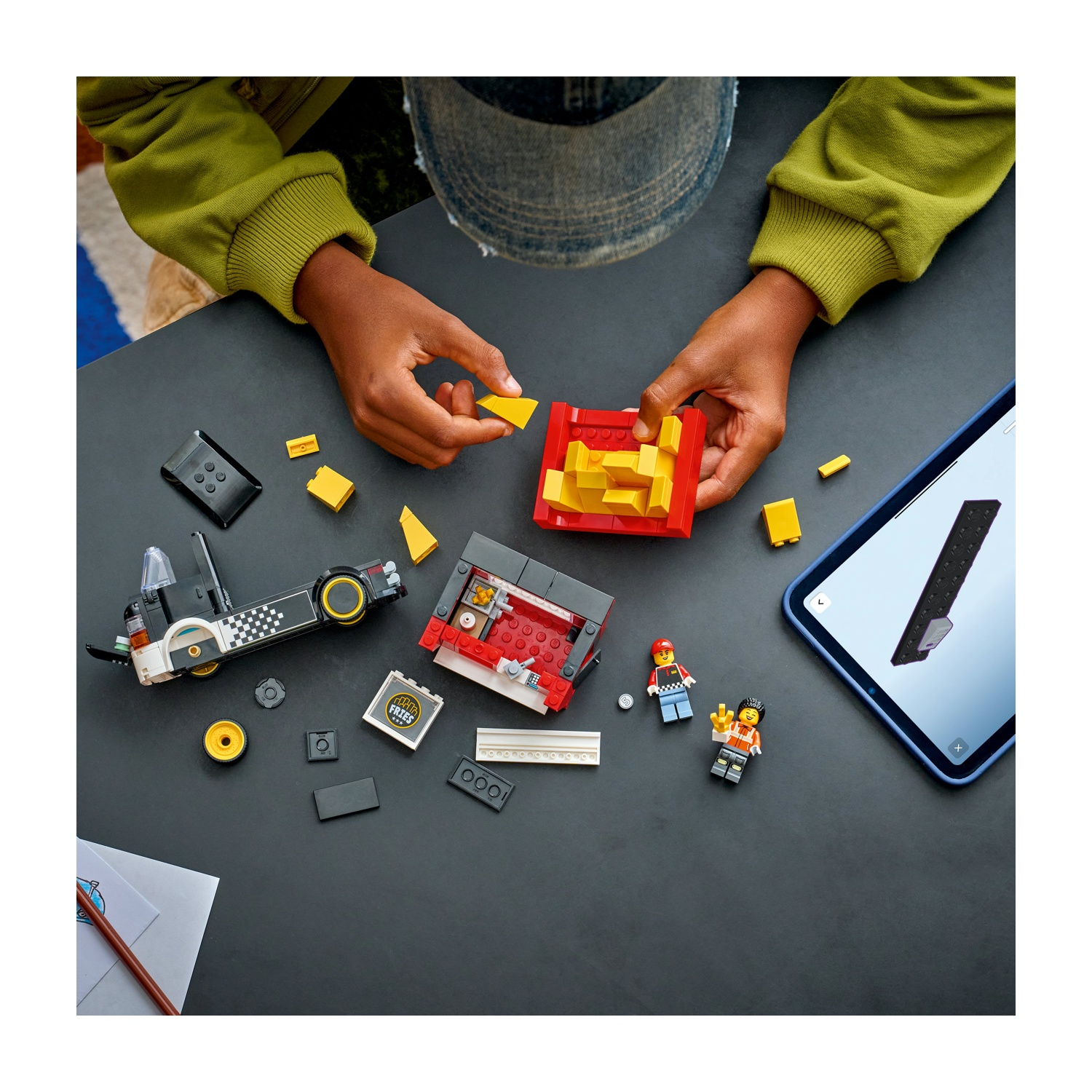 Mani che assemblano blocchi Lego su una superficie nera. Auto Lego, figure e tablet con istruzioni visibili.
