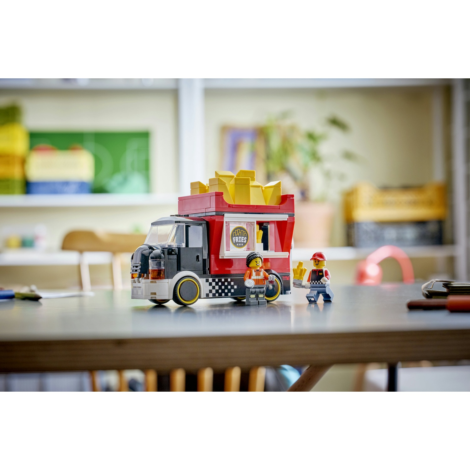 Food truck Lego con patatine fritte sopra. Due personaggi Lego sono in piedi accanto ad esso. Lo sfondo ha scaffali e una lavagna verde.