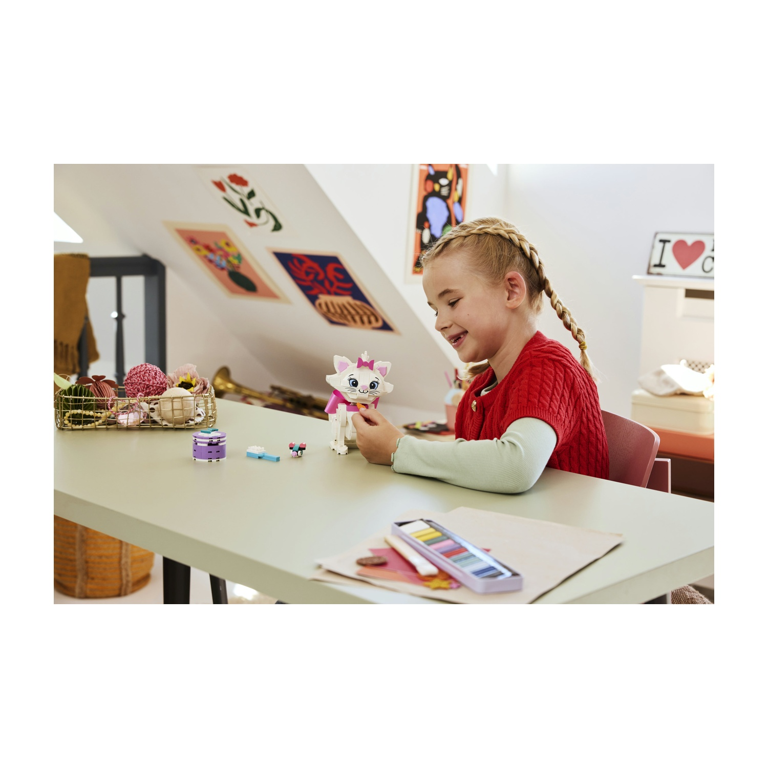 Una ragazza con le trecce gioca con un gatto giocattolo. Un tavolo contiene materiali artistici e un piccolo cestino. Opere d'arte sono appese al muro.