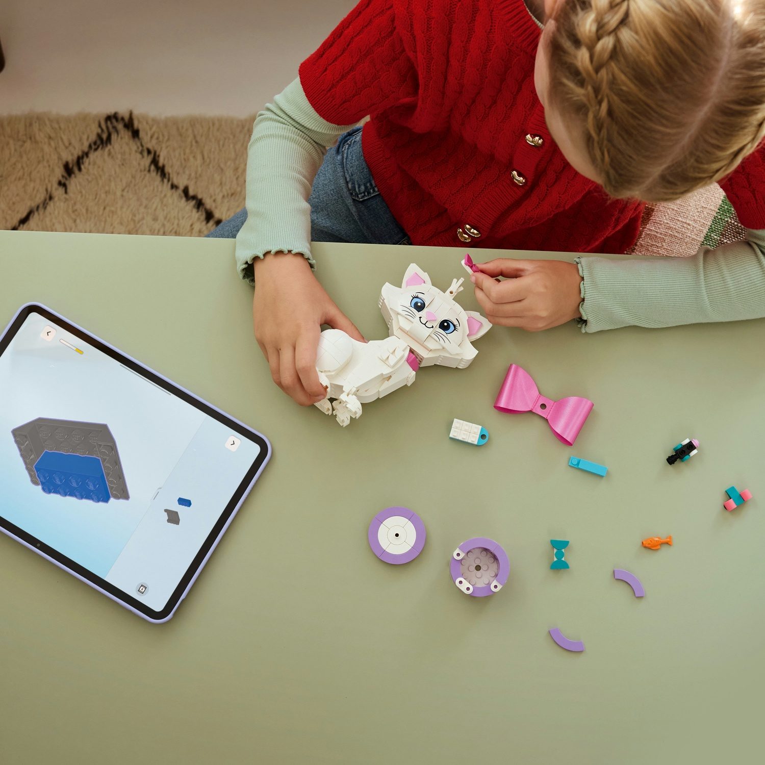 Una ragazza assembla un gatto Lego Marie. Un tablet mostra le istruzioni. Vari pezzi Lego e un fiocco rosa sono su un tavolo verde.