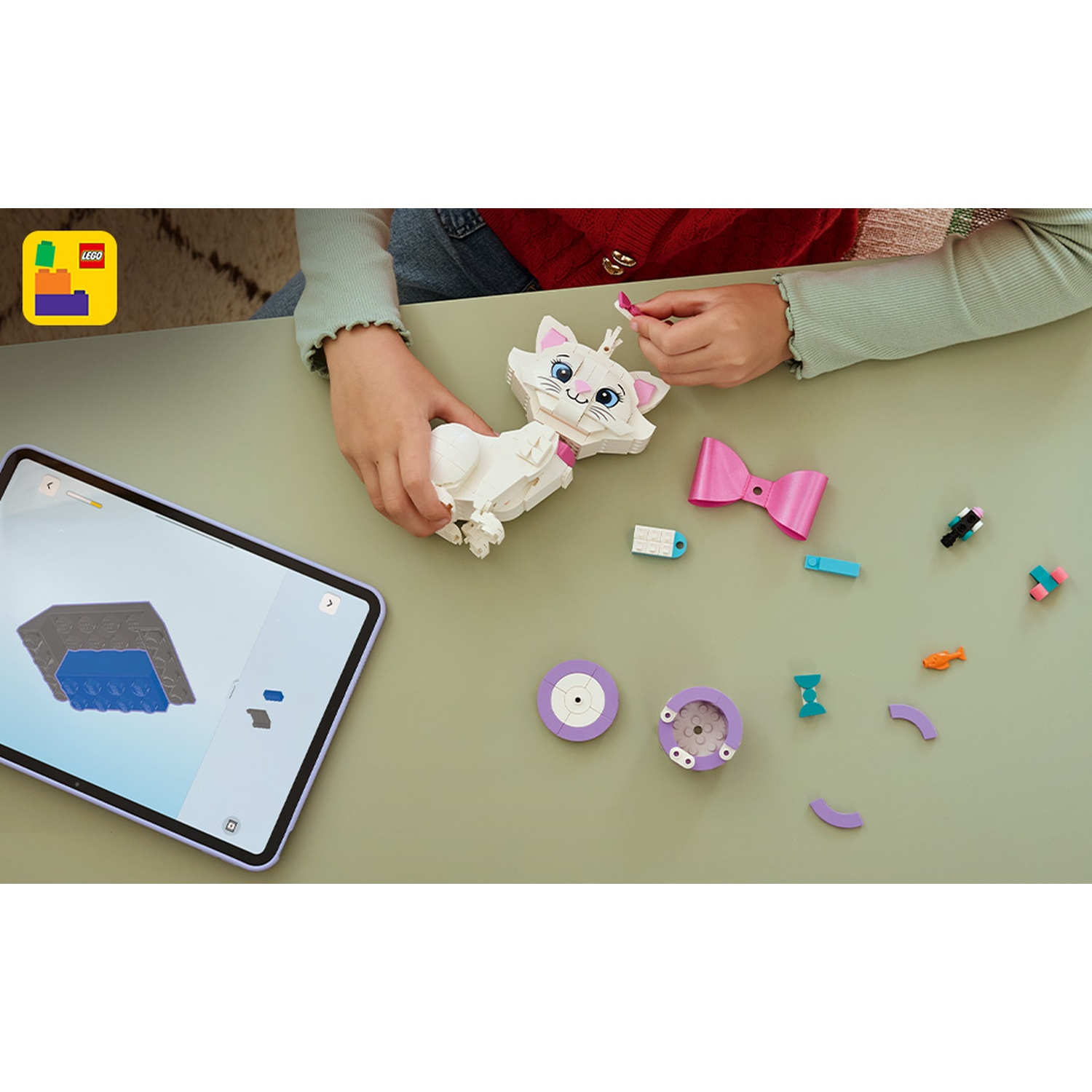Persona assembla gatto Lego bianco con tablet con istruzioni. Fiocco rosa, altri pezzi Lego su tavolo verde.