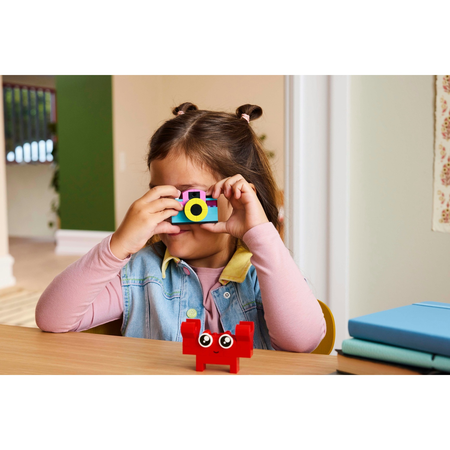 Una bambina guarda attraverso una macchina fotografica giocattolo. Un granchio di mattoni rossi è sul tavolo. Libri a destra.