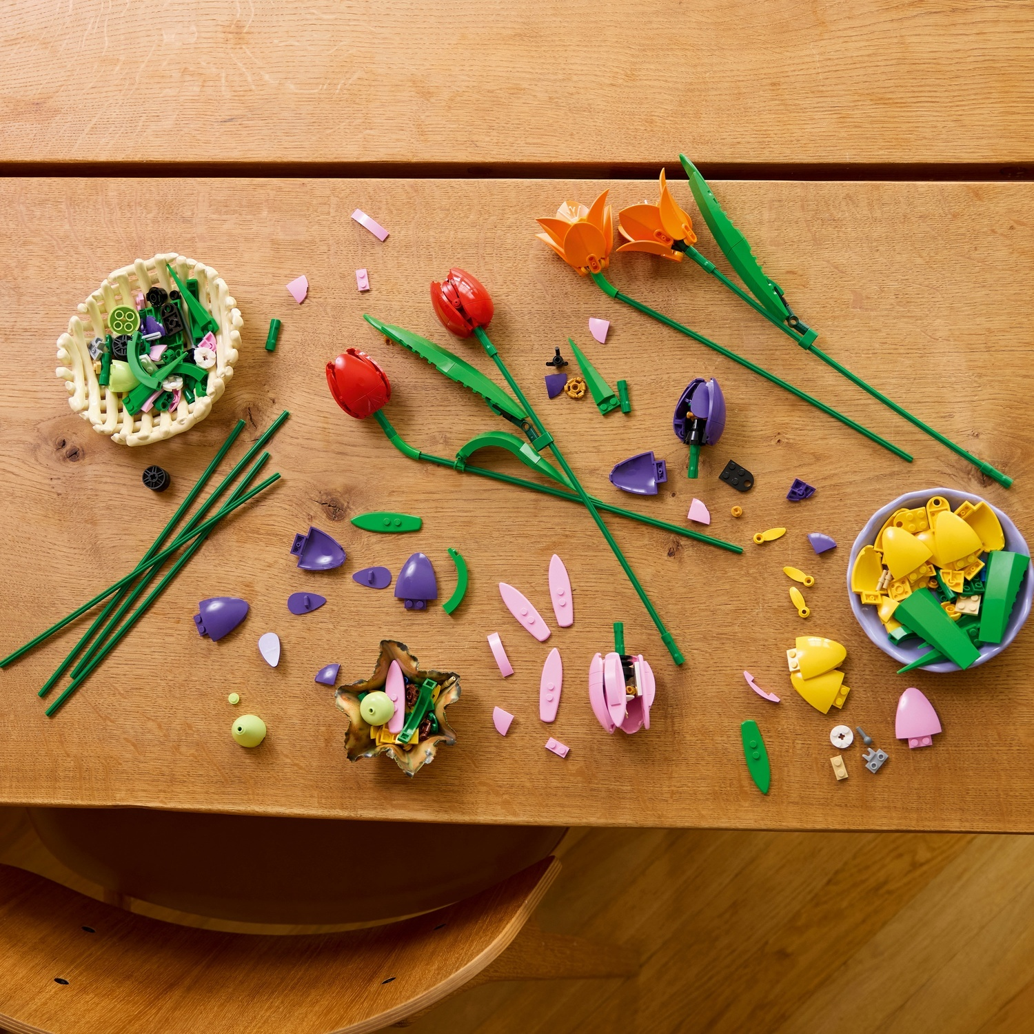 Tavolo di legno con pezzi di fiori Lego. Include steli verdi, petali rossi, arancioni e viola in cestini e sparsi.