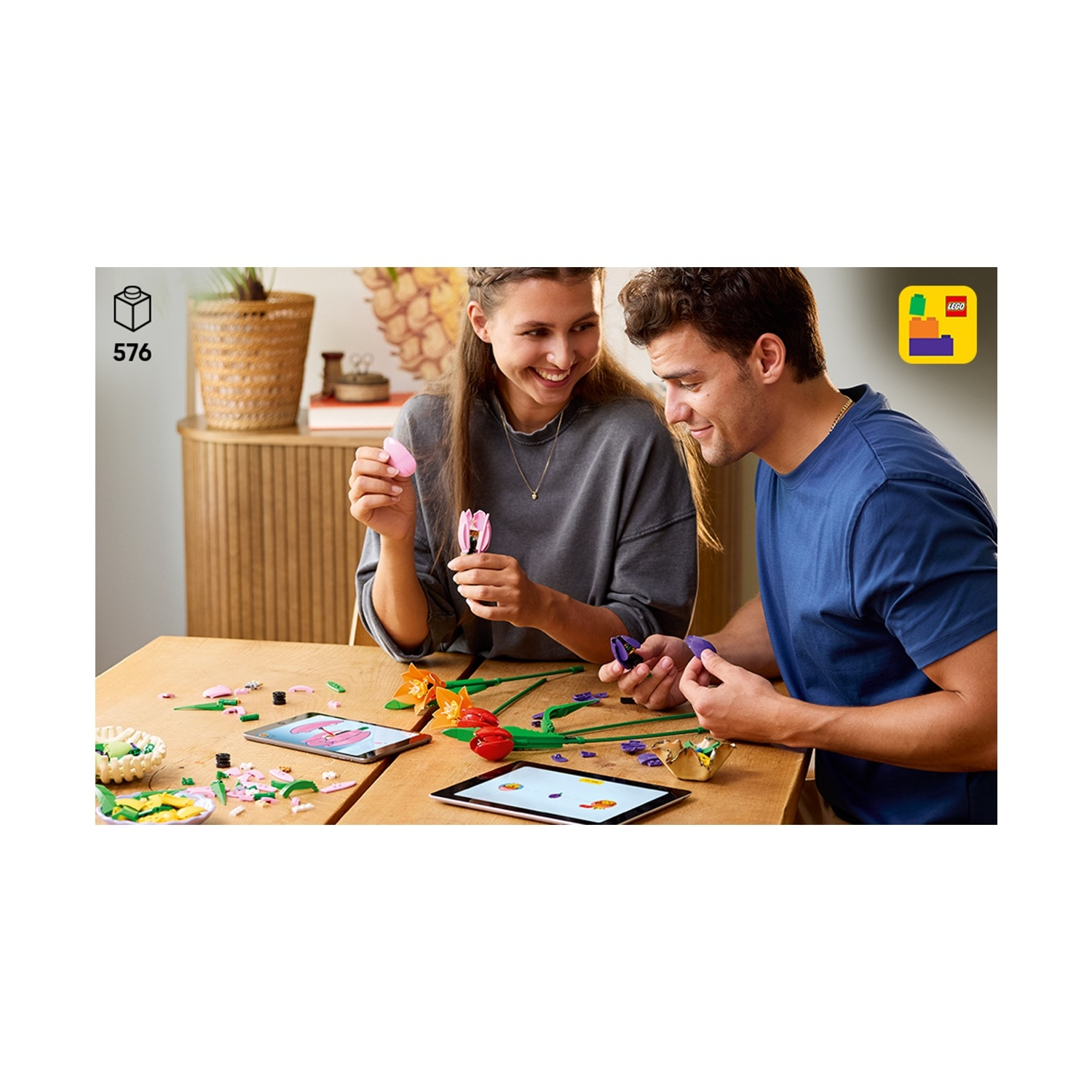 Una coppia assembla fiori LEGO a un tavolo. I tablet mostrano le istruzioni. Si stanno realizzando fiori rosa, arancioni e rossi.