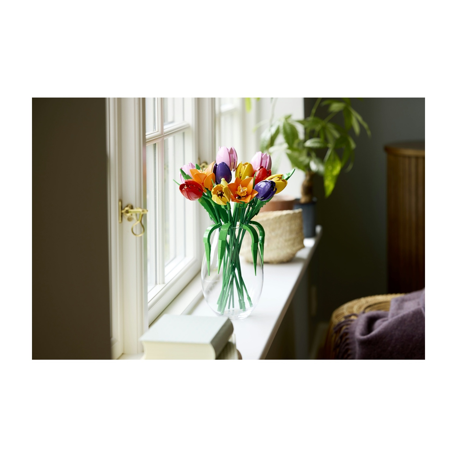 Un vaso di tulipani colorati si trova su un davanzale. Un libro, una pianta e una sedia in vimini con una coperta viola sono visibili.