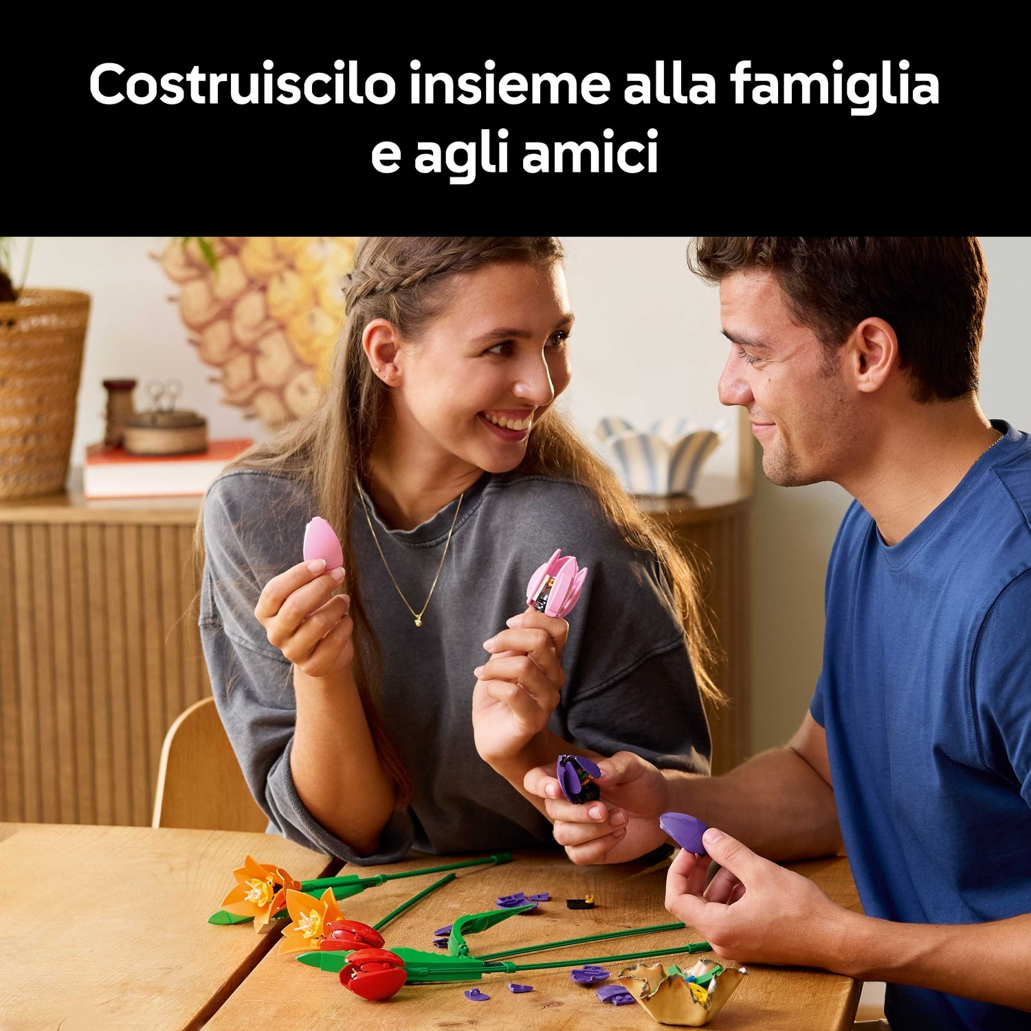 Un uomo e una donna assemblano fiori Lego a un tavolo. La donna tiene un tulipano rosa, sorridendo. L'uomo ha petali viola.