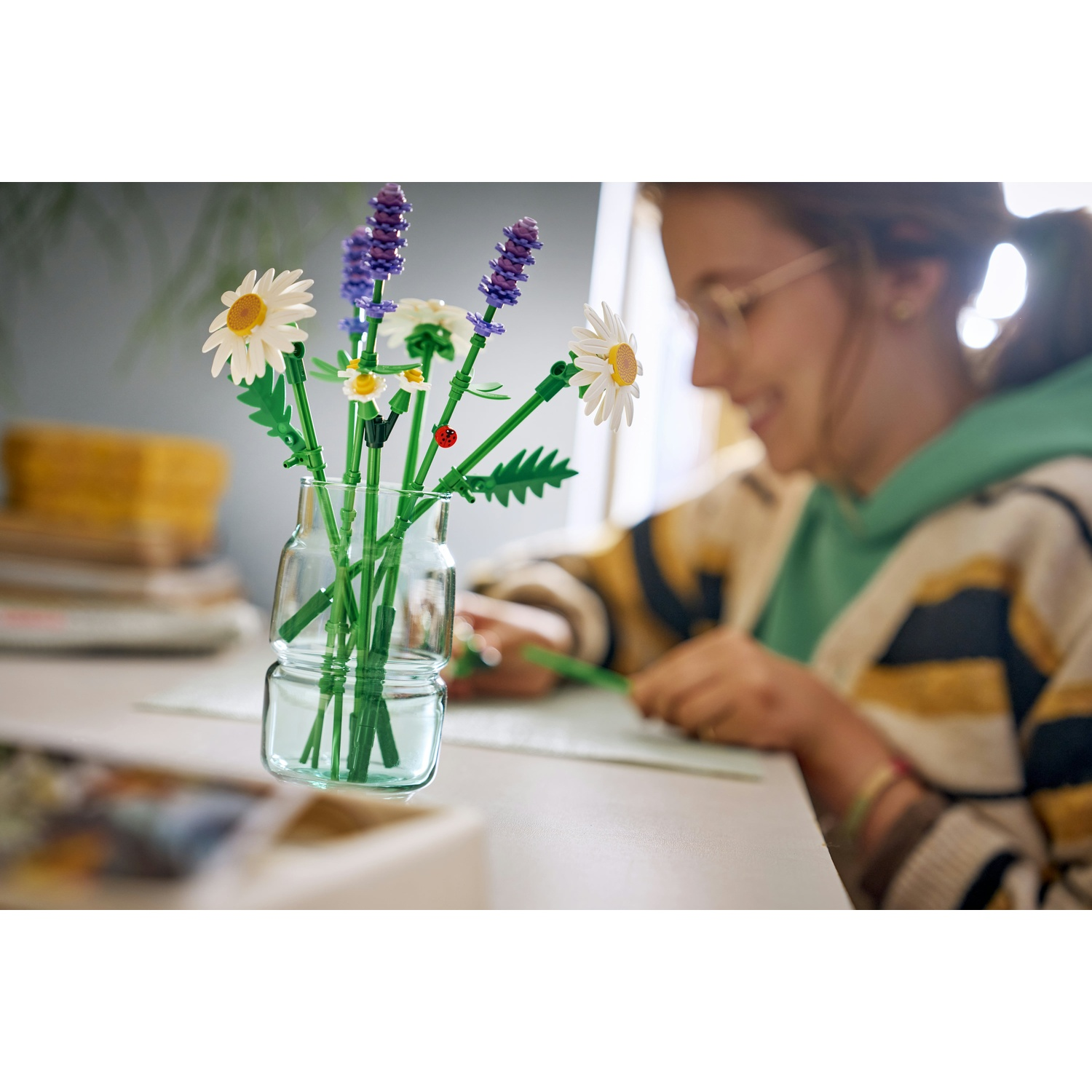 Un vaso di vetro con fiori, tra cui margherite e lavanda, su un tavolo. Una persona scrive sullo sfondo.