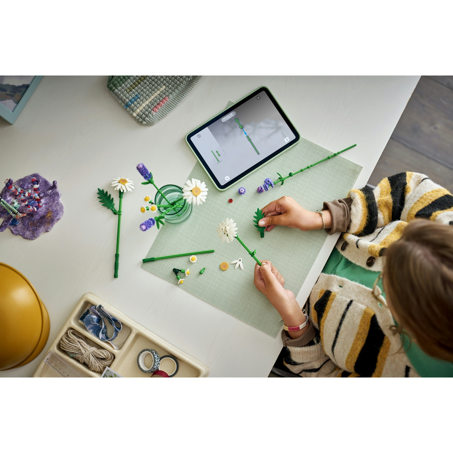 Una persona assembla steli di fiori su un tavolo. Un tablet mostra le istruzioni. Sono visibili margherite, strumenti e accessori.