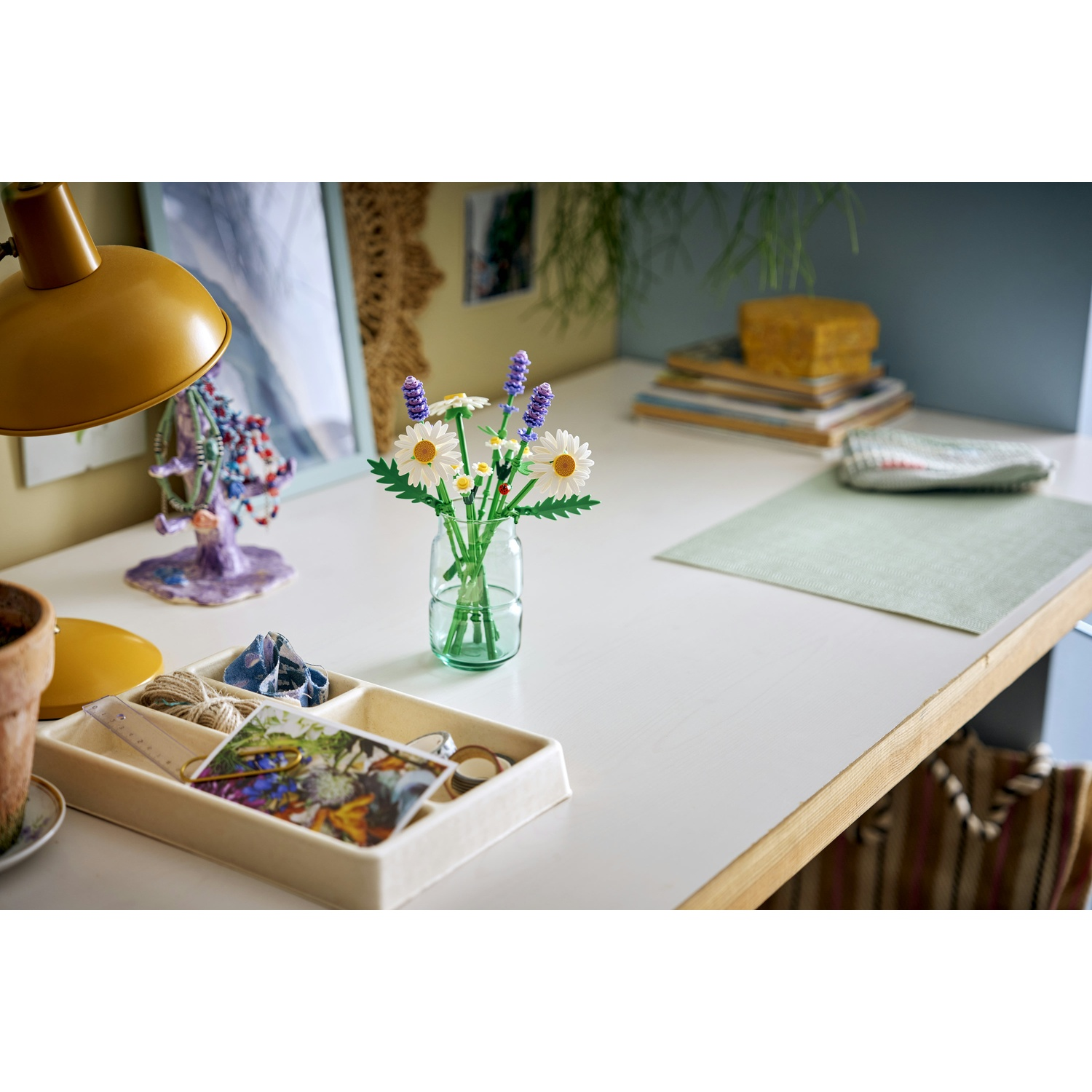 Una scrivania con un vaso di fiori, una lampada e un vassoio con oggetti. Sullo sfondo, una pila di libri e una borsa beige.