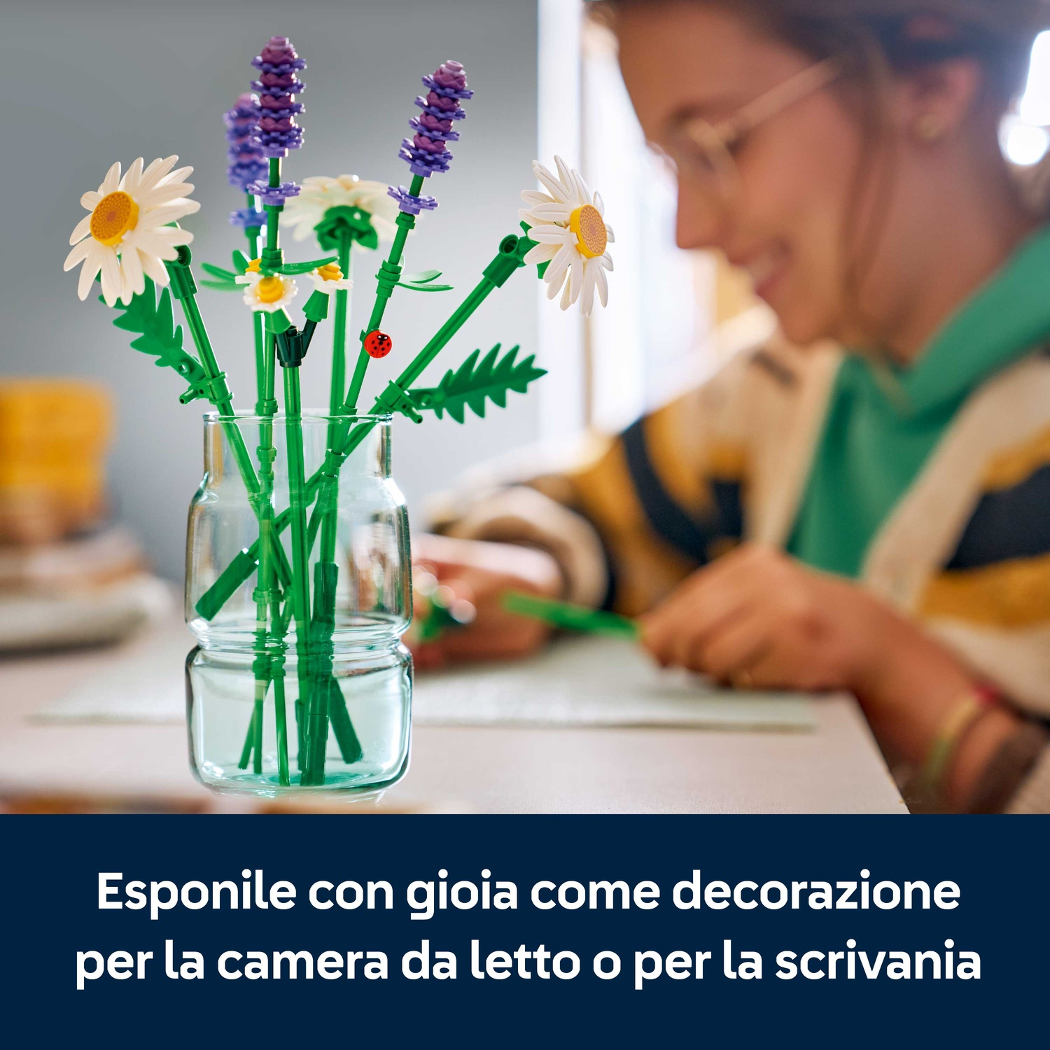 Un vaso con fiori Lego, tra cui margherite e lavanda, su un tavolo. Una persona è sullo sfondo, sfocata, che scrive.