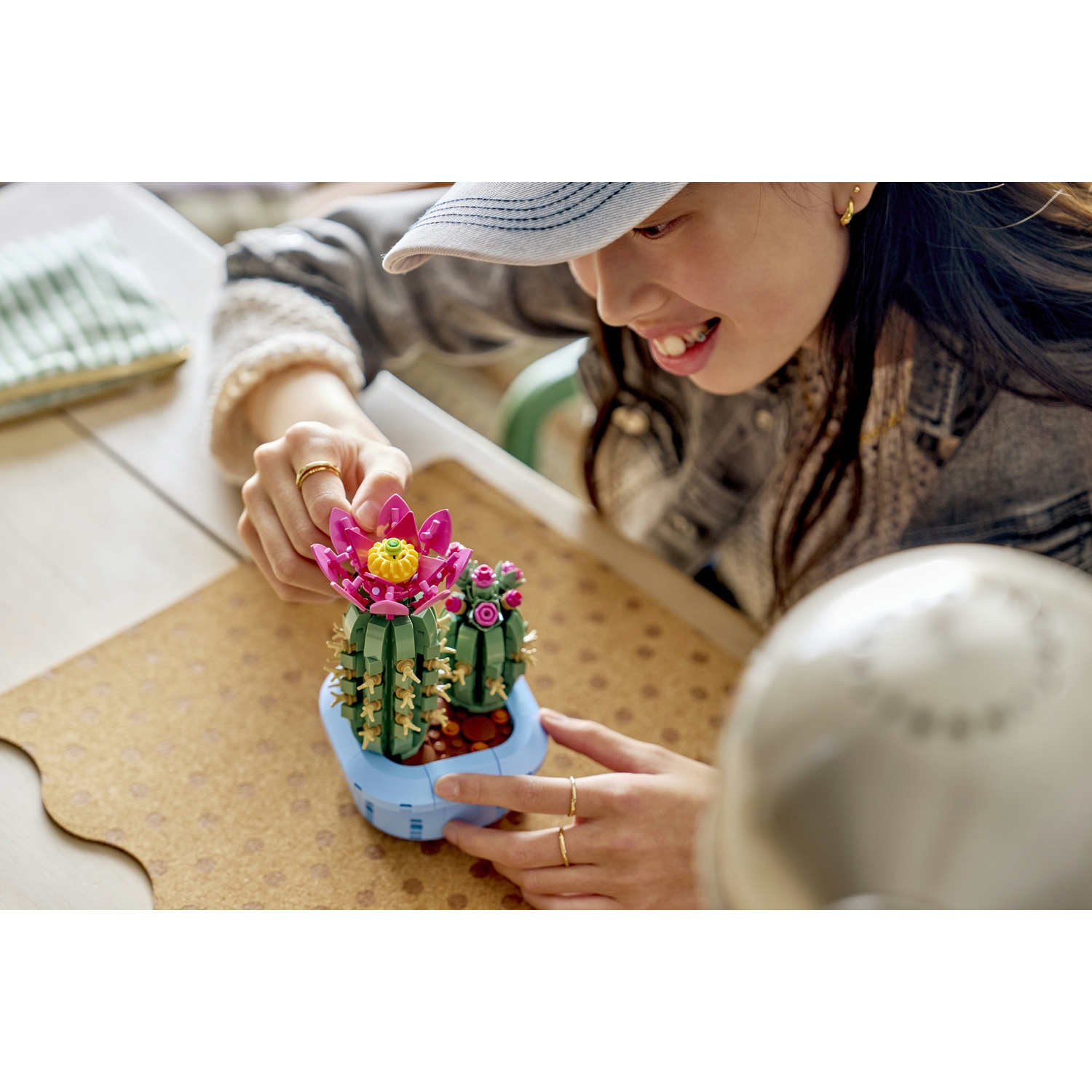 Una persona sorridente con un cappello tiene in mano un cactus Lego. Il cactus ha fiori rosa ed è in un vaso blu su un piano di sughero.