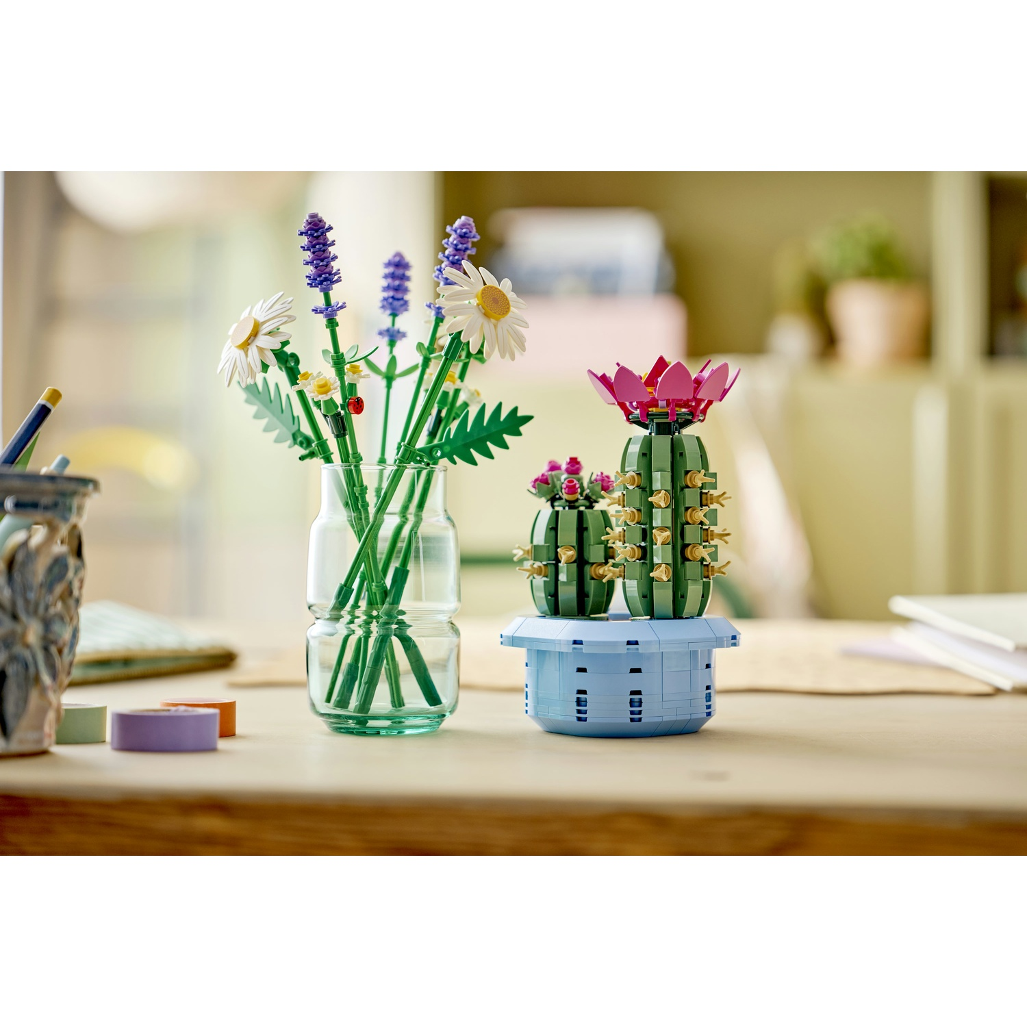 Un set Lego di cactus e fiori su un tavolo di legno. Un vaso di vetro contiene fiori Lego. Un vaso blu contiene il cactus.