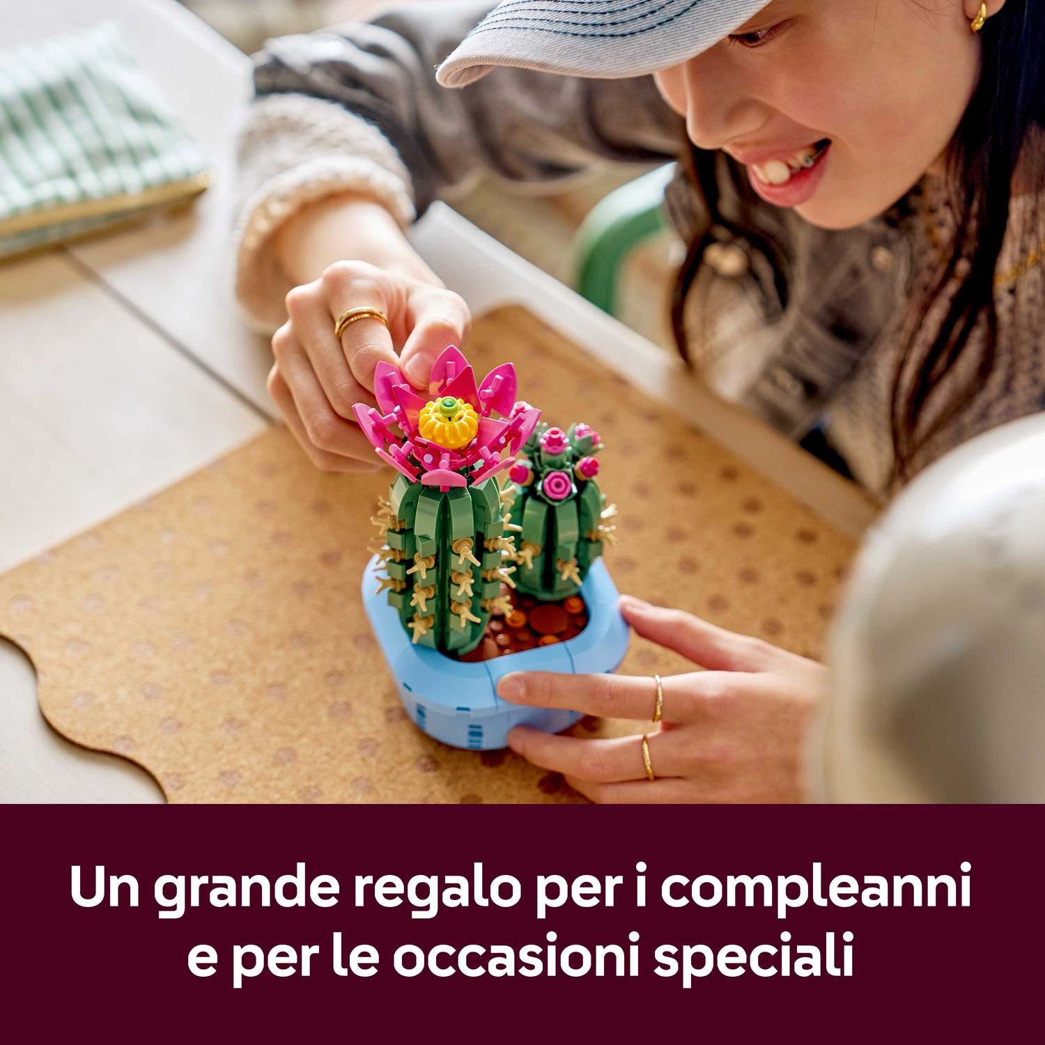 Una persona tiene in mano un cactus Lego in un vaso blu. Il cactus ha fiori rosa ed è su una superficie di legno. Testo in basso.