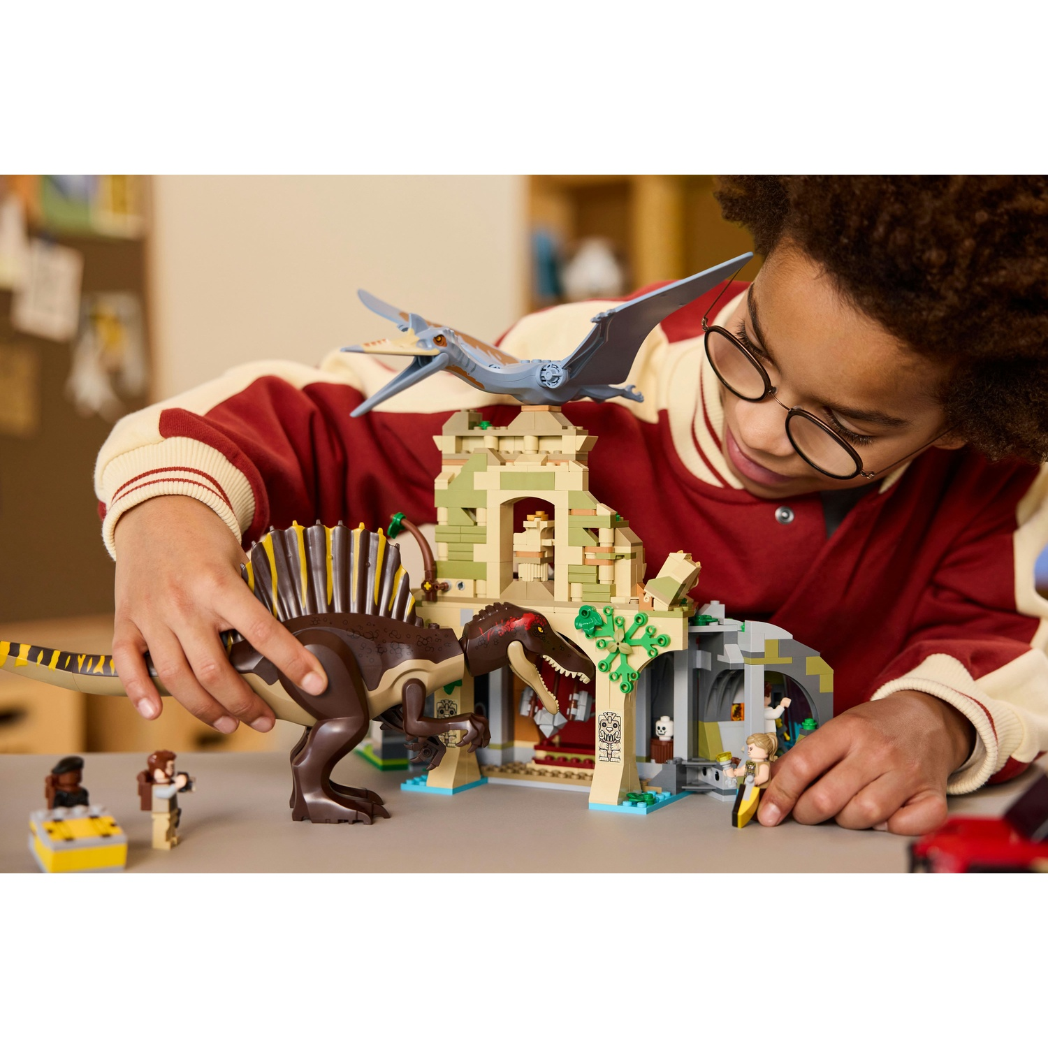 Un bambino con gli occhiali gioca con un set Lego, tra cui un dinosauro, un edificio e delle figure. Lo sfondo mostra una giacca rossa.