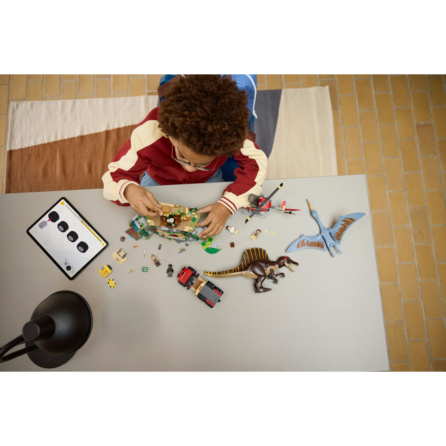 Un bambino gioca con i set Lego di dinosauri su un tavolo. Un tablet e una lampada sono sul tavolo. Sullo sfondo è visibile un tappeto.