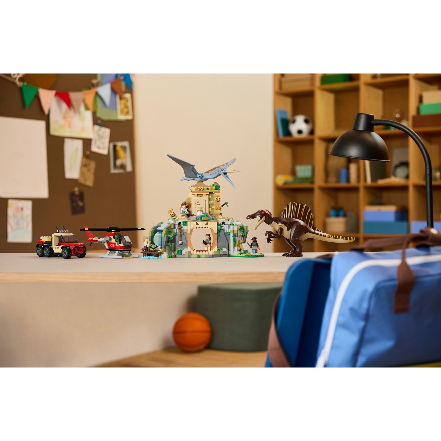 Una scrivania con giocattoli di dinosauri e veicoli. Sono presenti anche una borsa blu, una lampada e un pallone da basket. C'è una bacheca sulla parete.