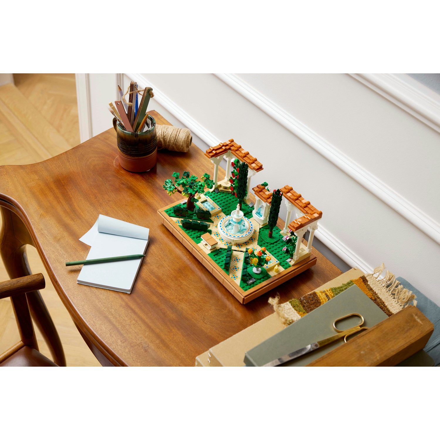 Un tavolo di legno mostra una scena di giardino Lego con una fontana. Un blocco per appunti e delle matite sono sul tavolo, accanto a una tazza di matite.