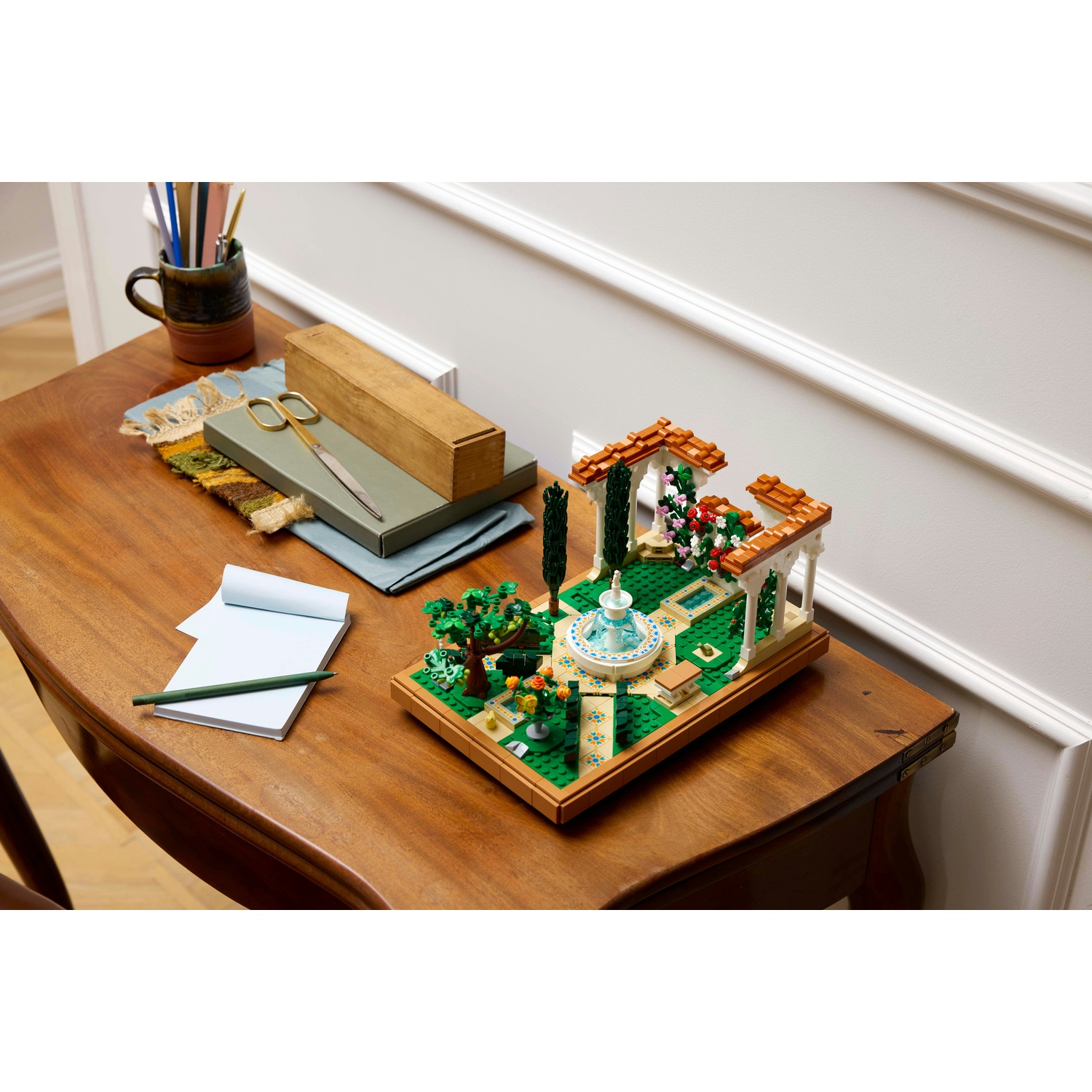 Un tavolo di legno mostra un set da giardino Lego, un blocco note, forbici e matite. Il giardino ha una fontana e fiori.