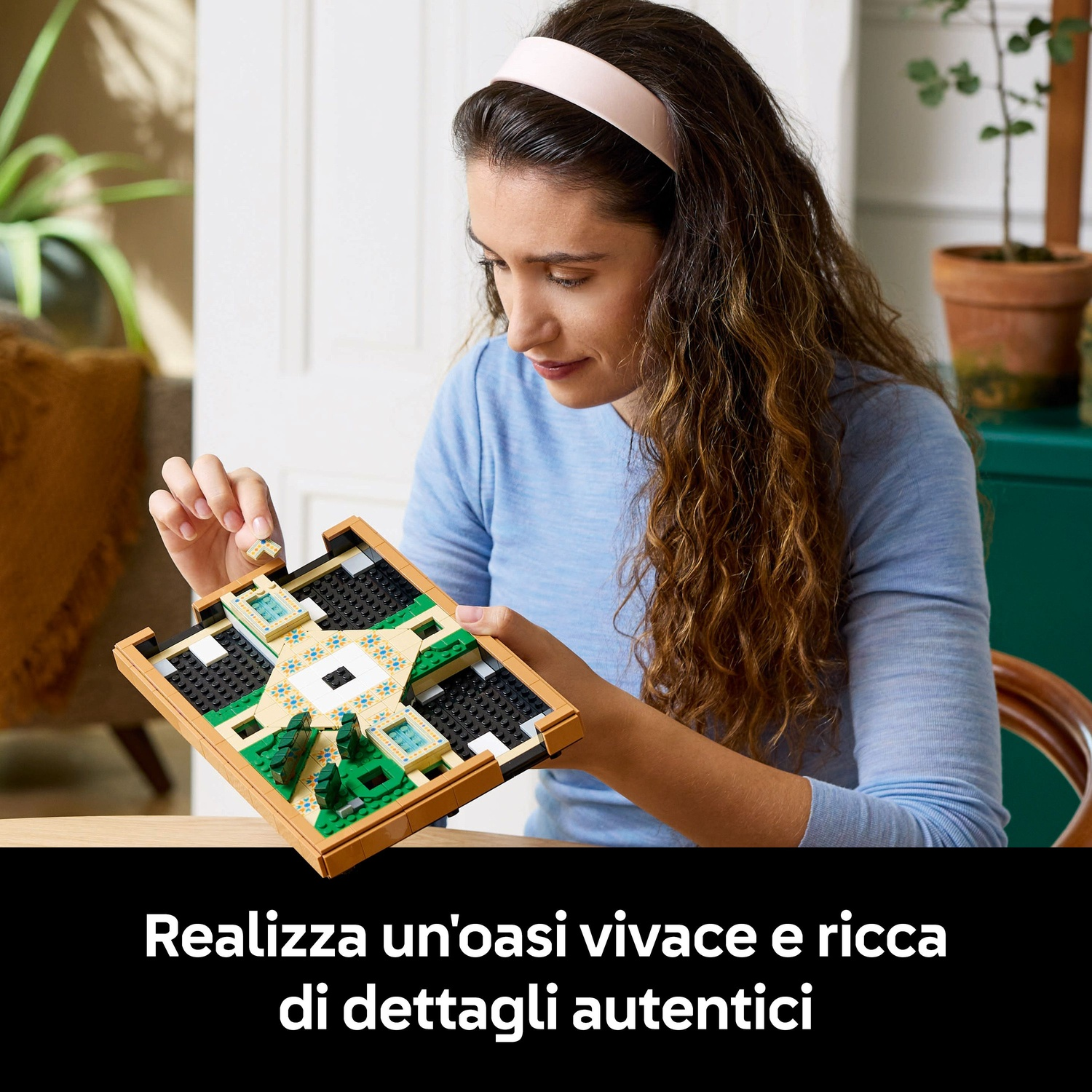 Una donna costruisce un modello Lego di un'oasi giardino. Indossa una camicia blu e una fascia rosa. Il modello include elementi verdi e beige.