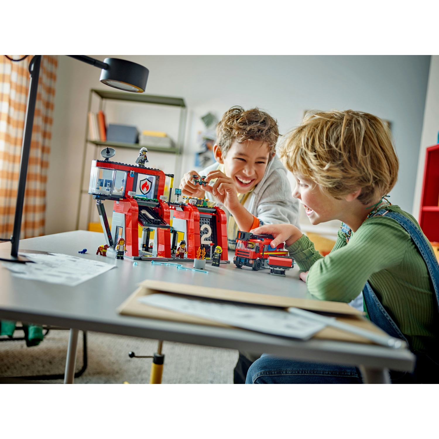 Due ragazzi giocano con un set della stazione dei pompieri Lego. Uno tiene un piccolo aereo, l'altro tocca un camion dei pompieri.