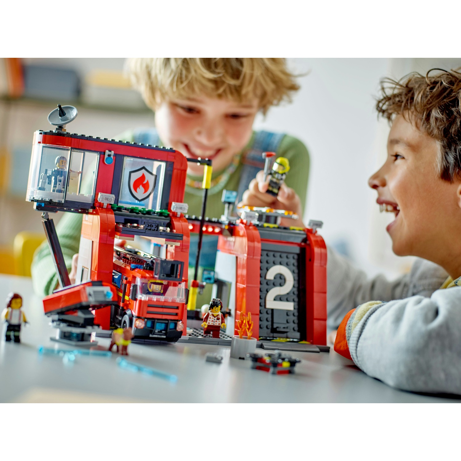 Due ragazzi giocano con una stazione dei pompieri Lego, uno tiene una figura. La stazione ha un camion dei pompieri rosso e il numero 2.