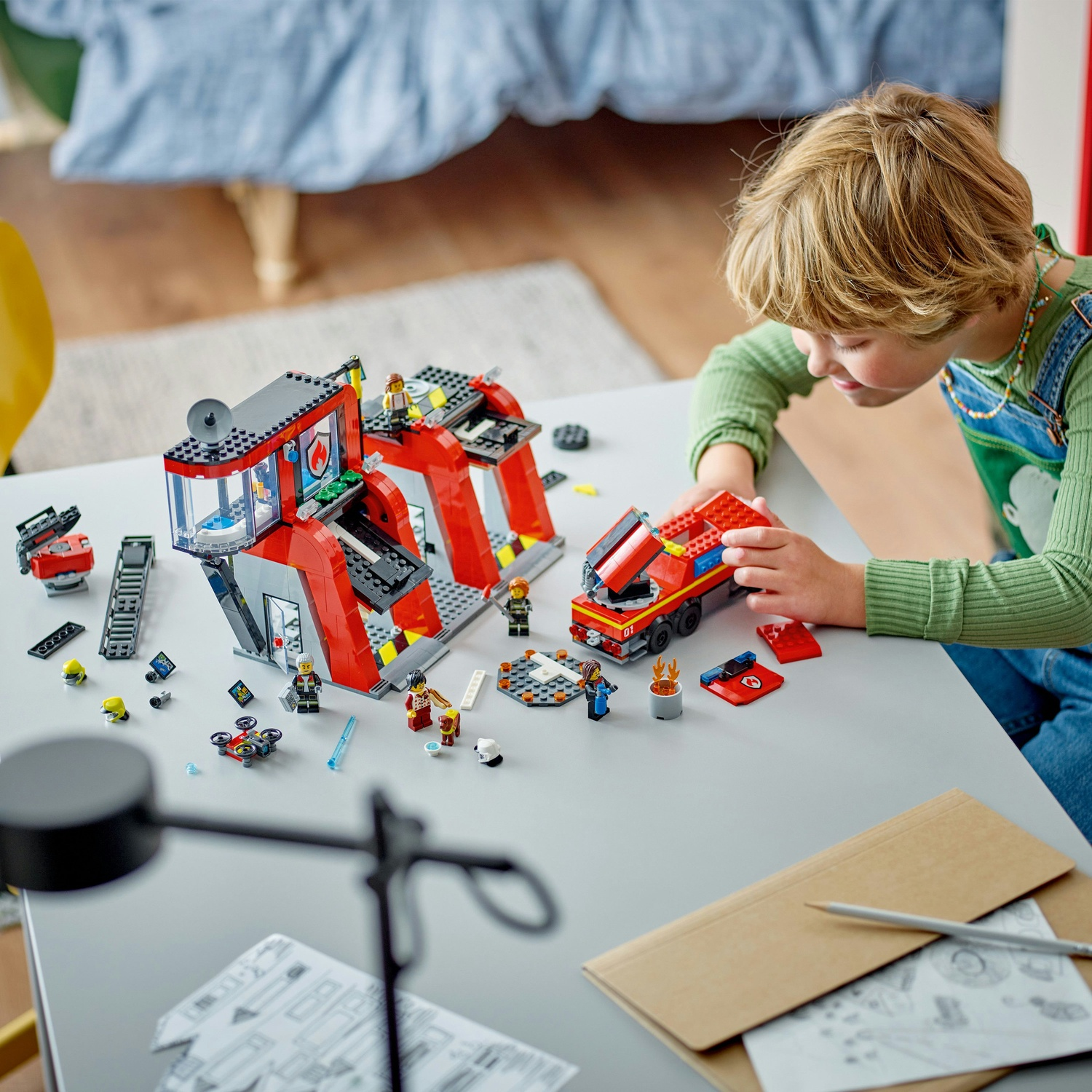 Un bambino gioca con una stazione dei pompieri Lego e un camion dei pompieri. La scena è su un tavolo grigio, con pezzi Lego sparsi.