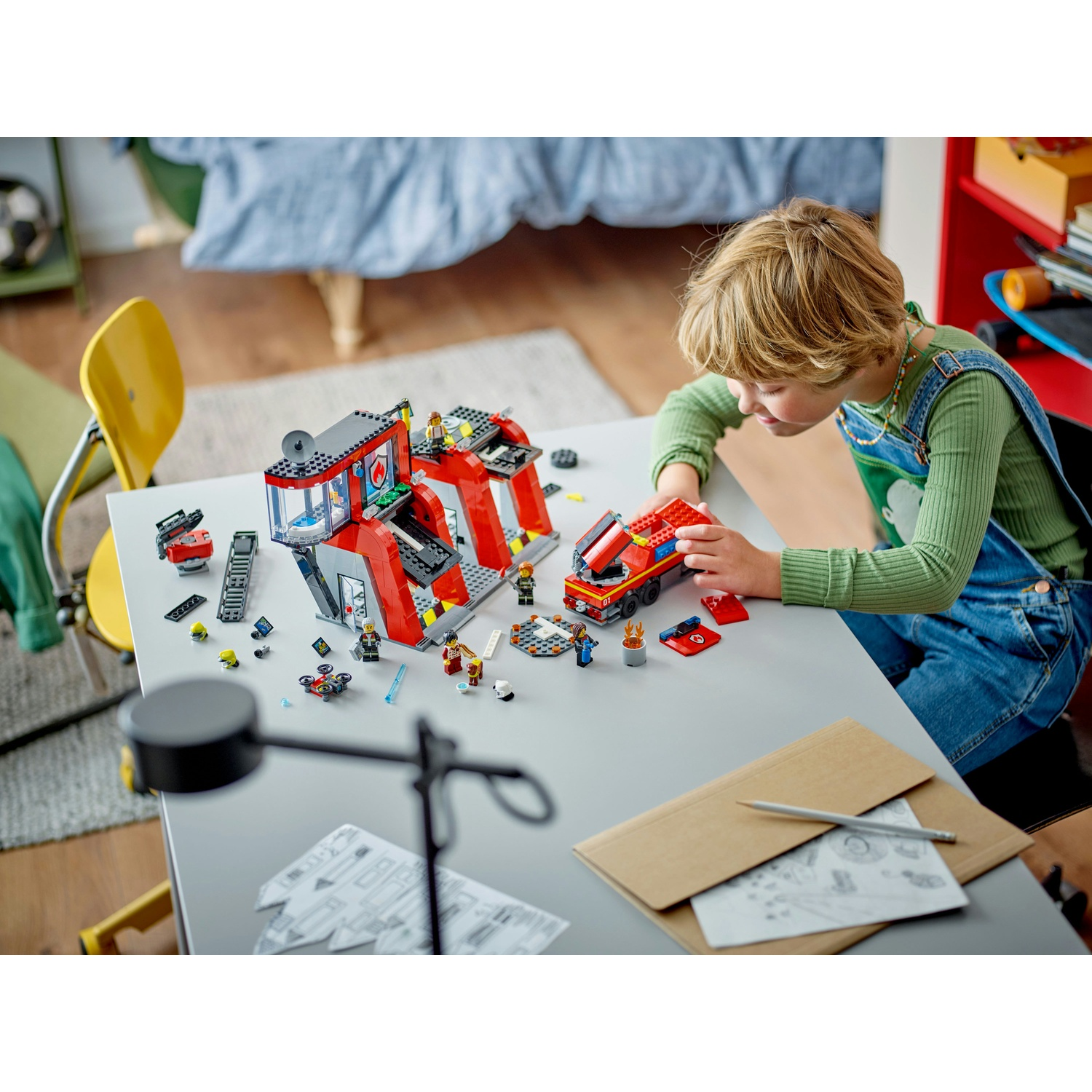 Un bambino costruisce una caserma dei pompieri e un camion Lego. La scena è ambientata su un tavolo grigio con altri pezzi Lego sparsi.