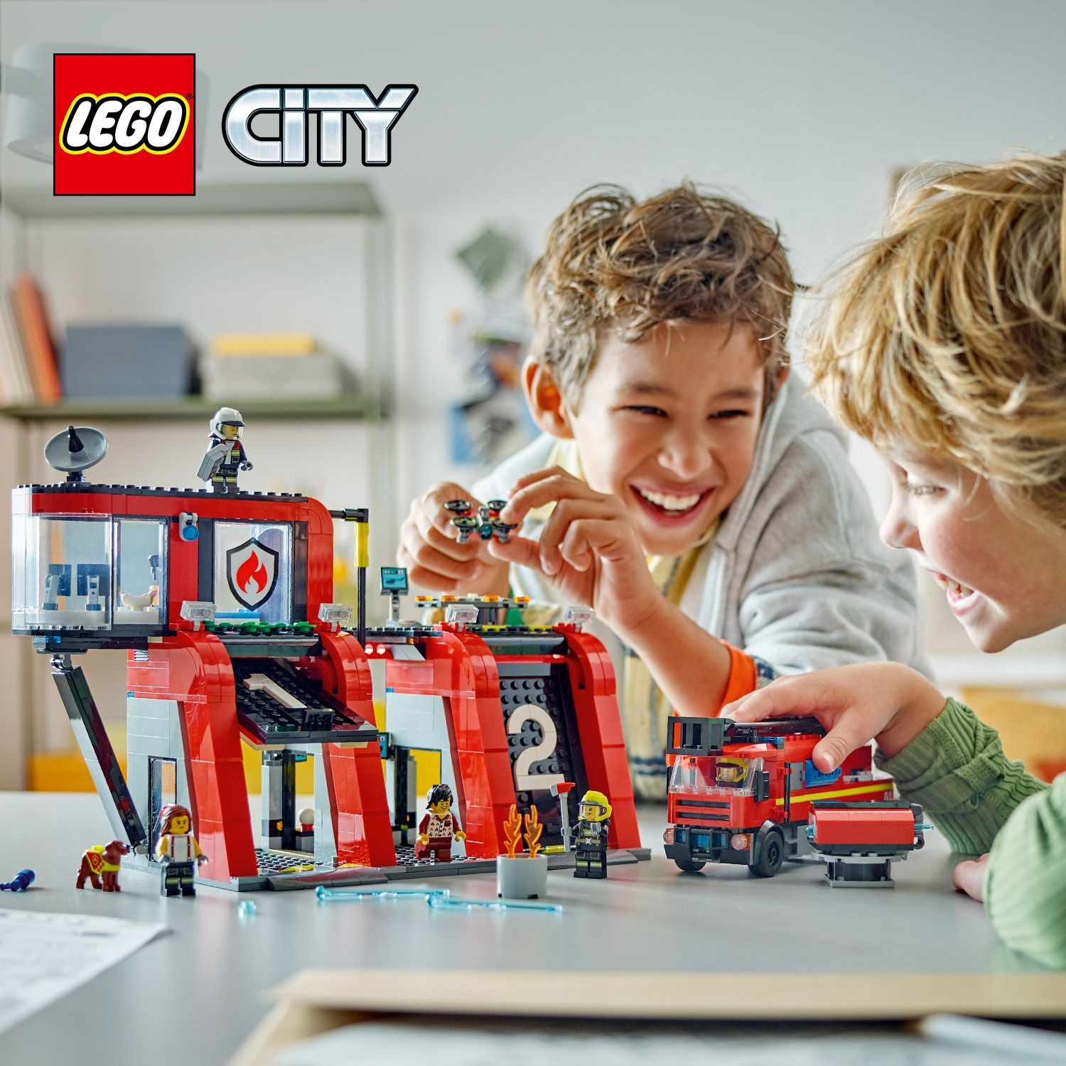 Due ragazzi giocano con un set LEGO City della stazione dei pompieri. Il set include un'autopompa, personaggi e un edificio con uno scudo.