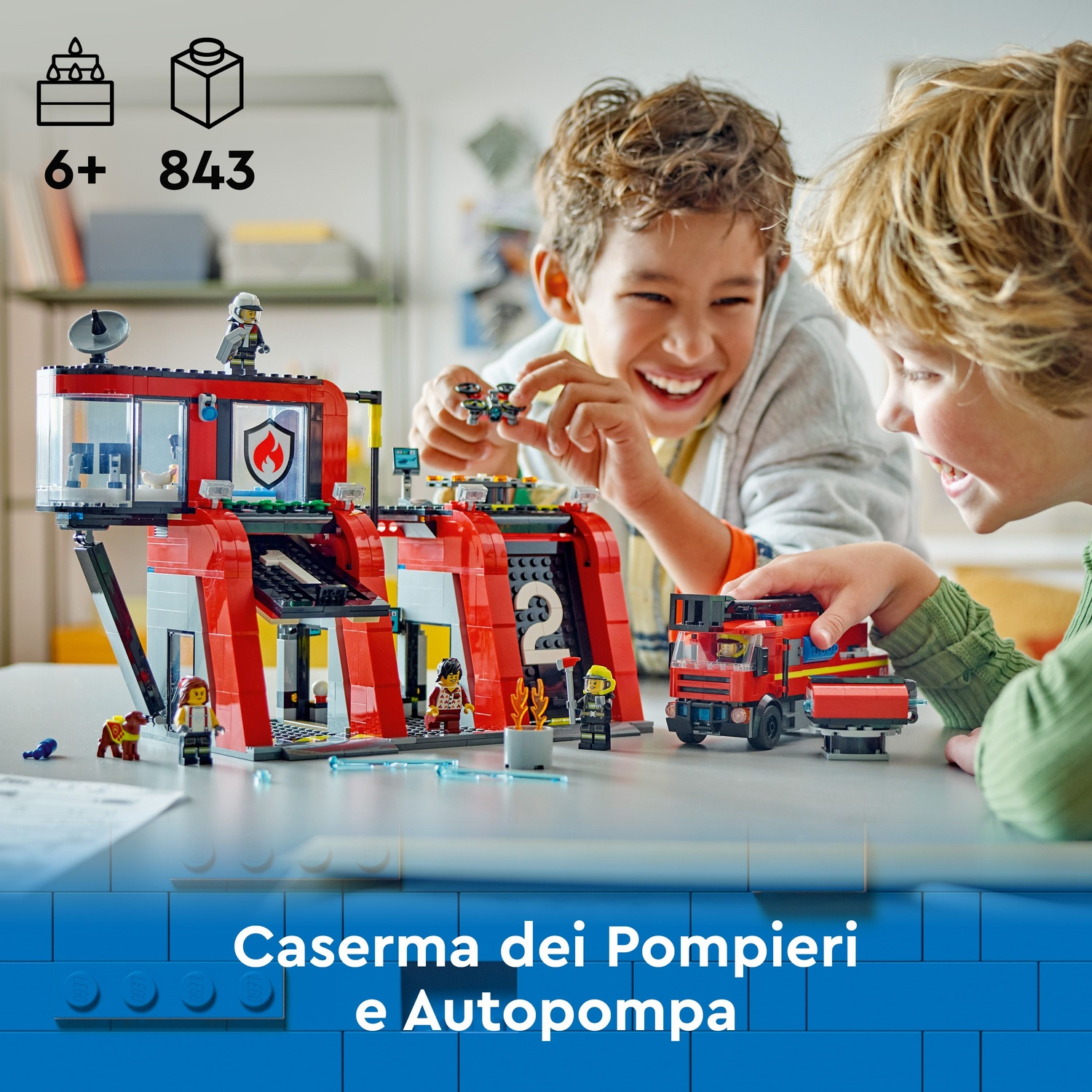 Due bambini che giocano con un set di caserma dei pompieri Lego. Il set include un camion dei pompieri e diverse figure Lego. Lo sfondo è un tavolo bianco.