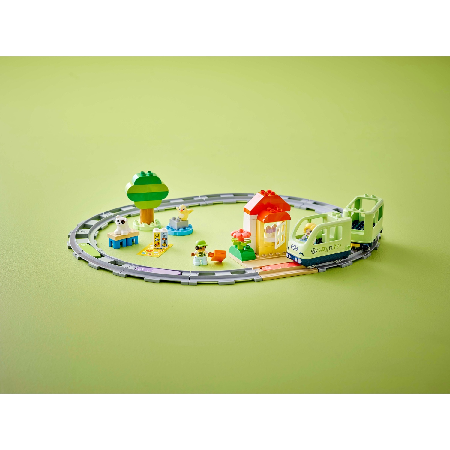 Un set di treni Lego su sfondo verde. Include un treno, una stazione, un albero e altre figure su un binario circolare.