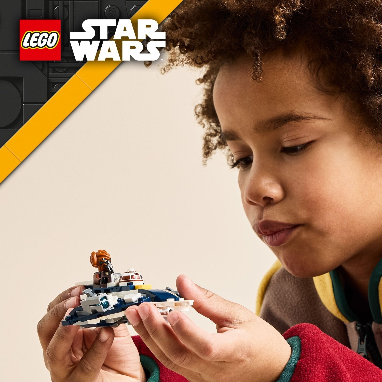 Un bambino tiene in mano una nave Lego di Star Wars. La nave è blu, bianca e grigia. Il bambino indossa una giacca rossa, marrone e gialla.