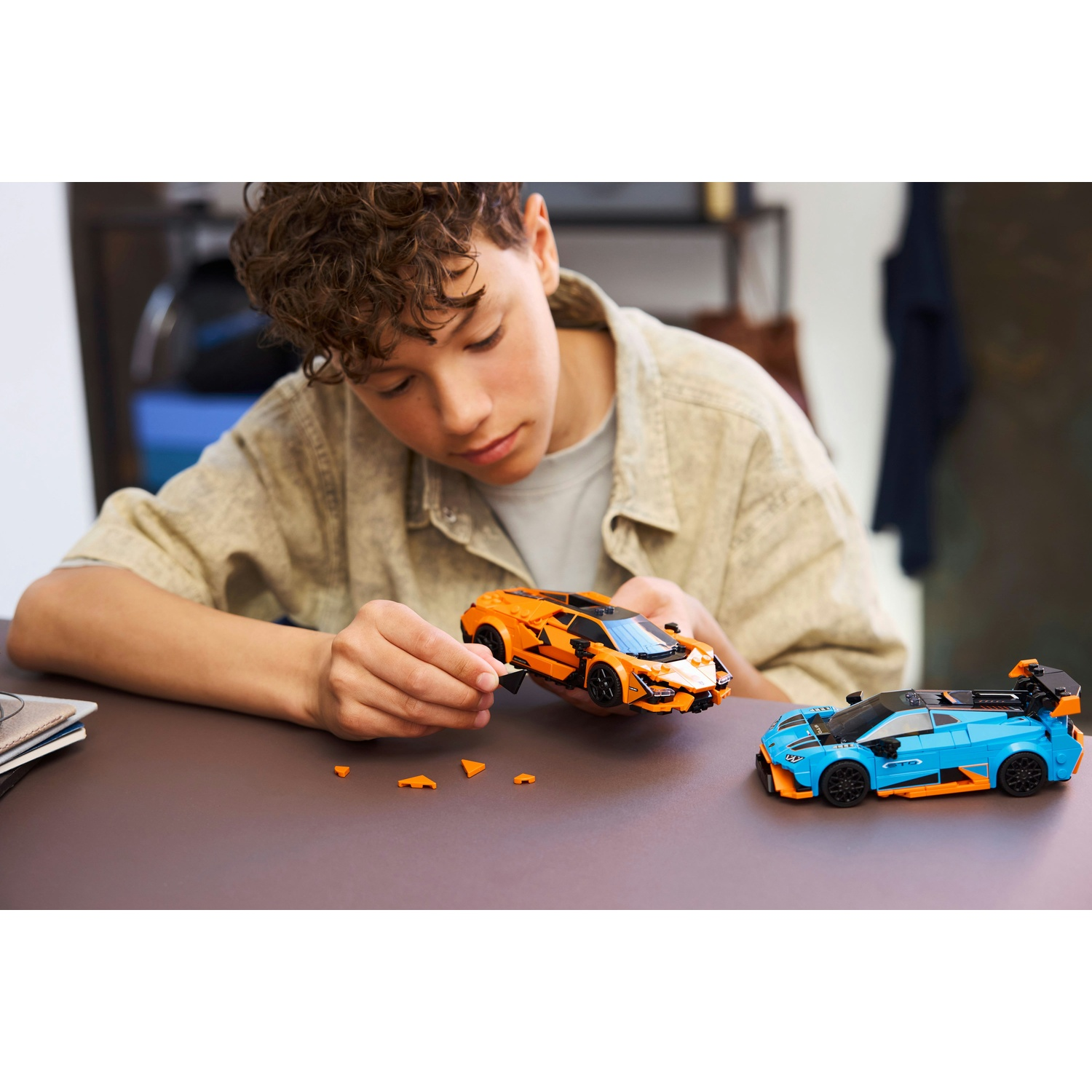 Un ragazzo assembla un'auto Lego arancione. Un'auto Lego blu è vicina. Parti sparse su un tavolo marrone.