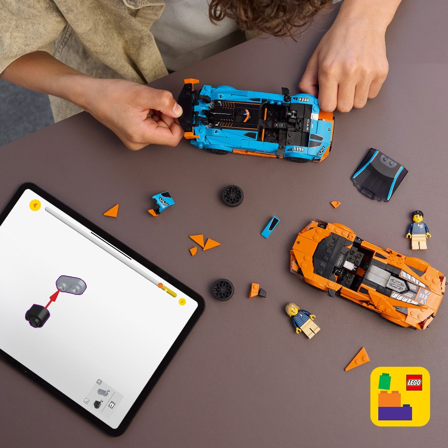 Mani che assemblano un'auto Lego blu e arancione. Un'auto Lego arancione, due personaggi Lego e un tablet con le istruzioni.