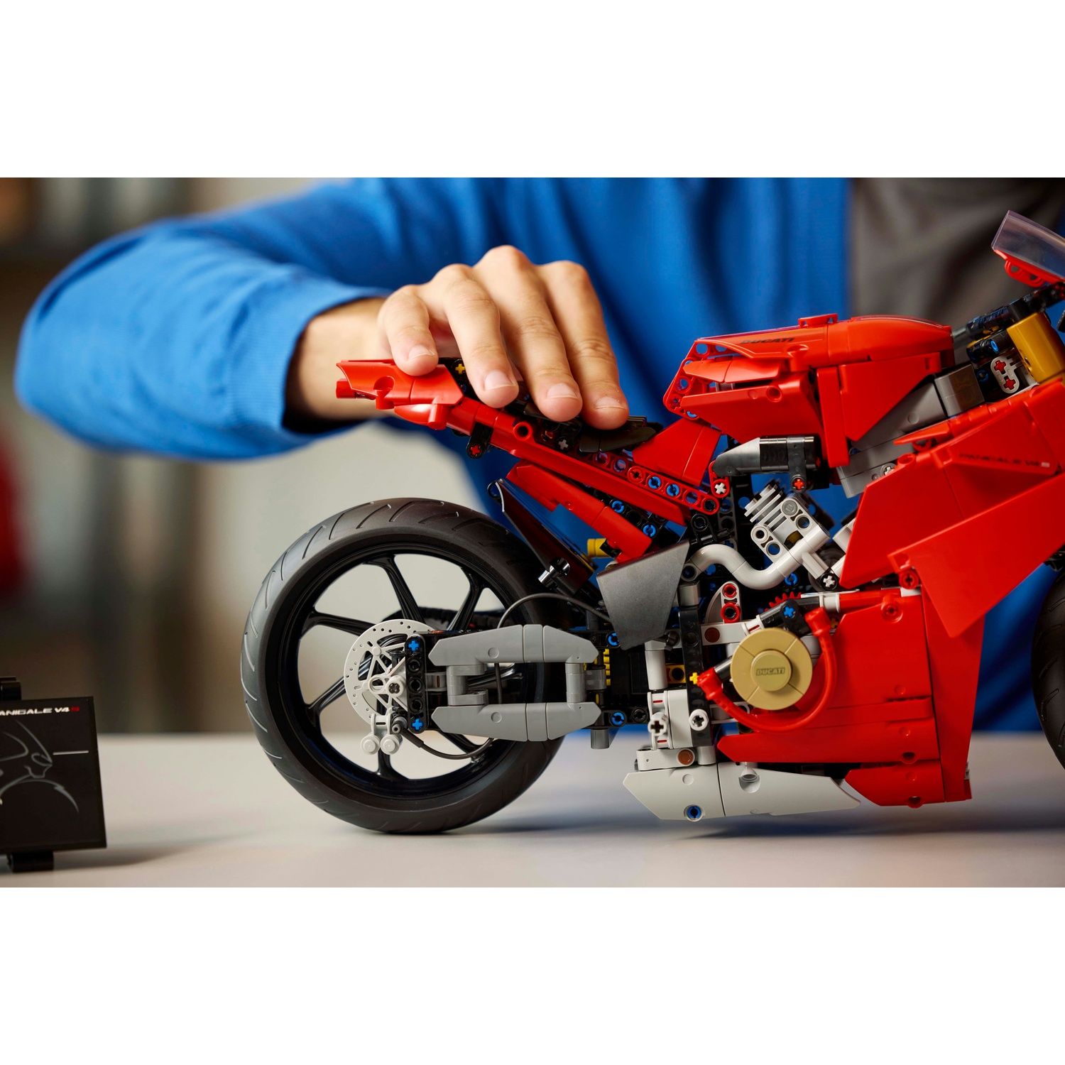 Una mano tocca una moto Lego rossa. La moto è su una superficie bianca con una scheda informativa nera a sinistra.