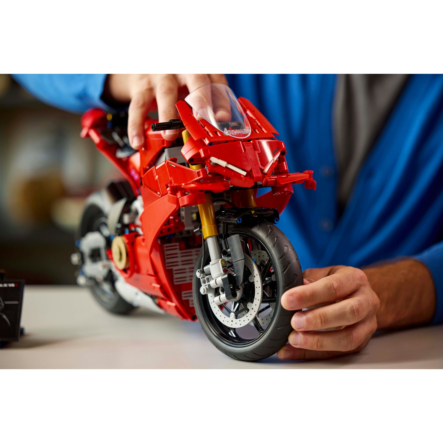 Una persona tiene in mano una moto giocattolo rossa su una superficie bianca. La moto ha pneumatici neri e un parabrezza trasparente. La persona indossa una camicia blu.