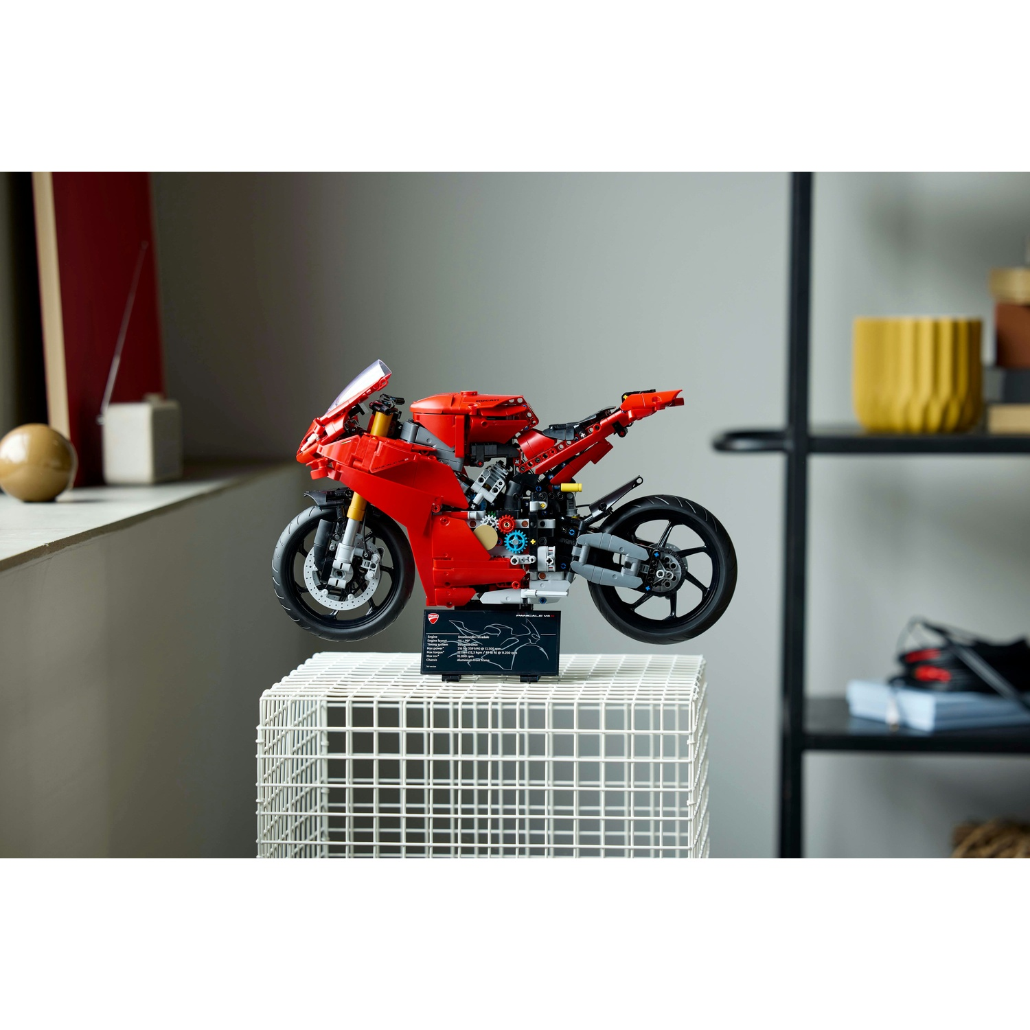 Un modello di moto Ducati rossa è esposto su un supporto a griglia bianca. Lo sfondo mostra una mensola con oggetti e una finestra.