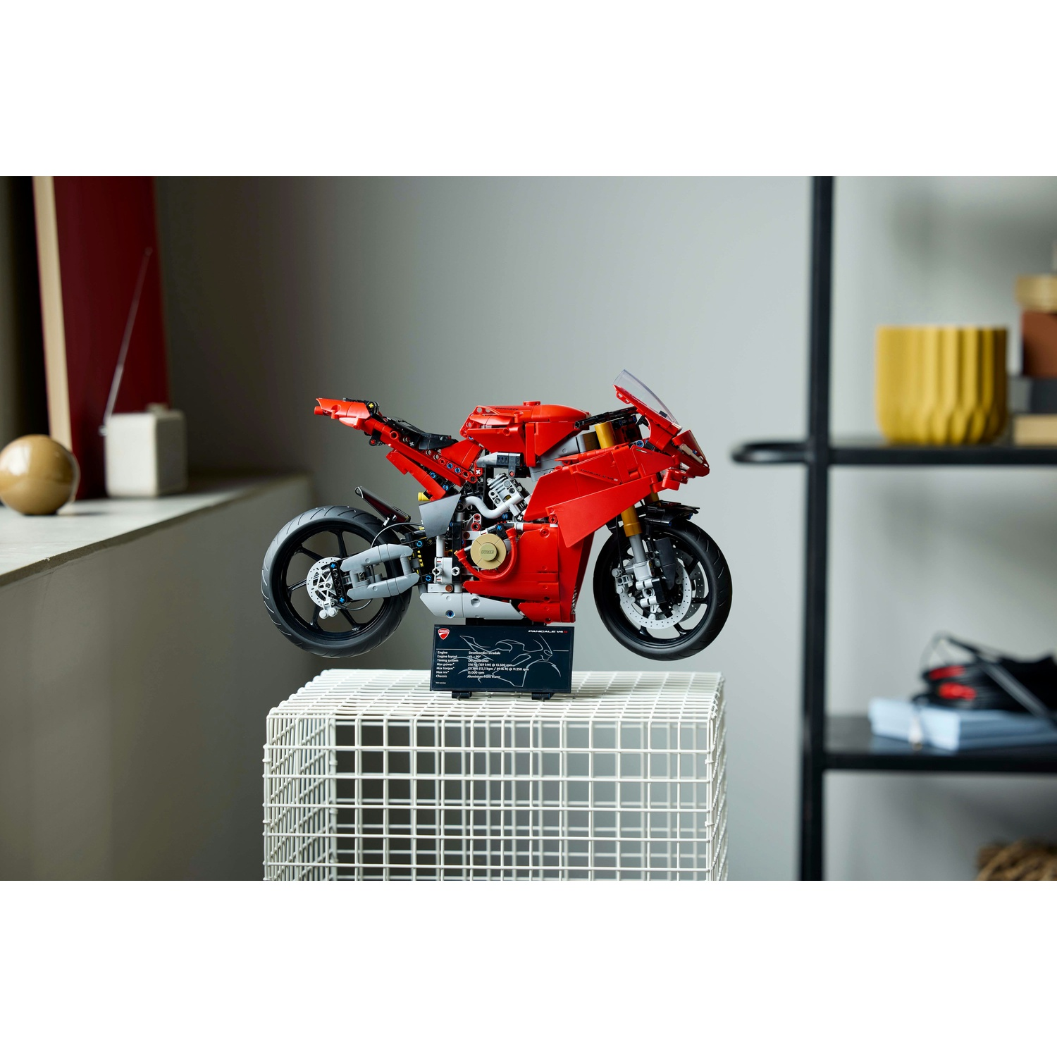 Una moto Ducati Lego rossa è esposta su un supporto di filo bianco. Sullo sfondo una mensola con decorazioni. Una finestra è a sinistra.