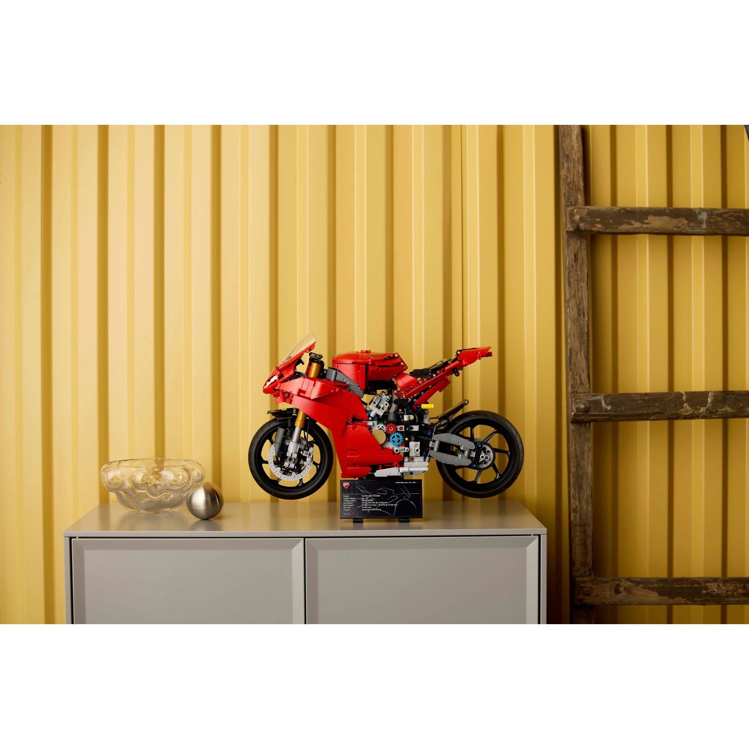 Modello di moto Lego rossa su un mobile grigio, con sfondo giallo ondulato e una scala di legno a destra.