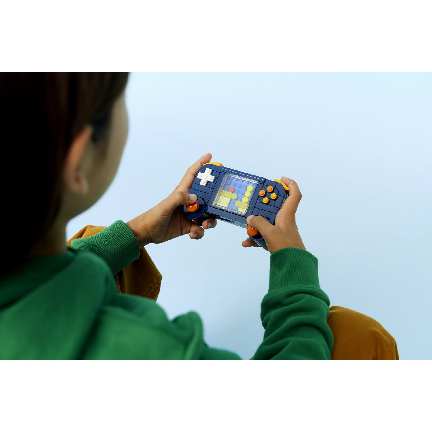 Un bambino tiene in mano una console di gioco portatile LEGO su uno sfondo azzurro. Il gioco mostra blocchi che cadono. Il bambino indossa una felpa con cappuccio verde.