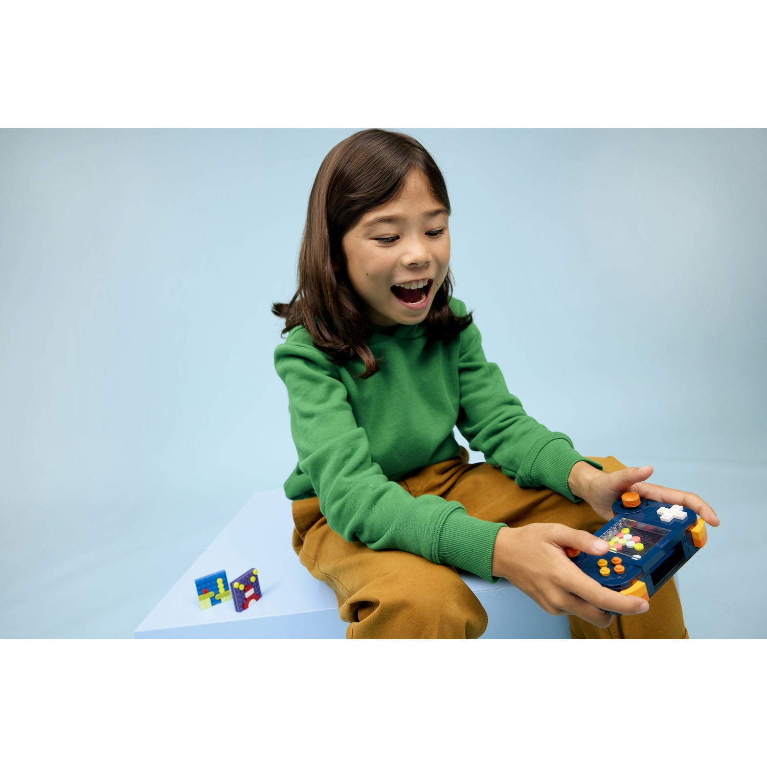 Un bambino con un maglione verde e pantaloni marroni gioca con un gioco portatile. Sono presenti una superficie e uno sfondo blu.