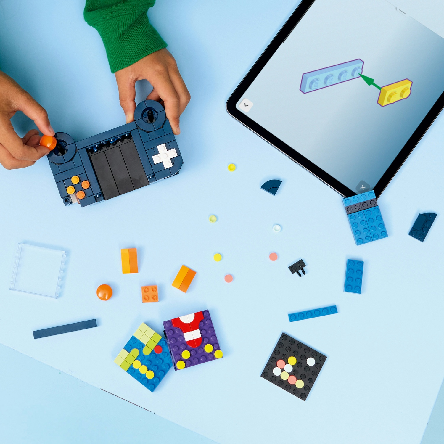Mani che assemblano un controller Lego blu con pulsanti arancioni. Un tablet mostra le istruzioni. Vari mattoncini Lego di diversi colori sparsi.