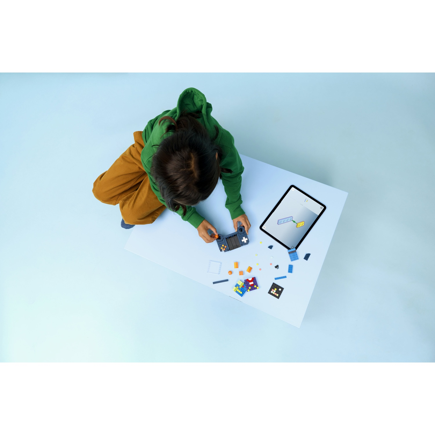 Un bambino con una felpa con cappuccio verde e pantaloni marroni gioca con i giocattoli su un tavolo bianco. C'è un tablet e vari blocchi colorati.