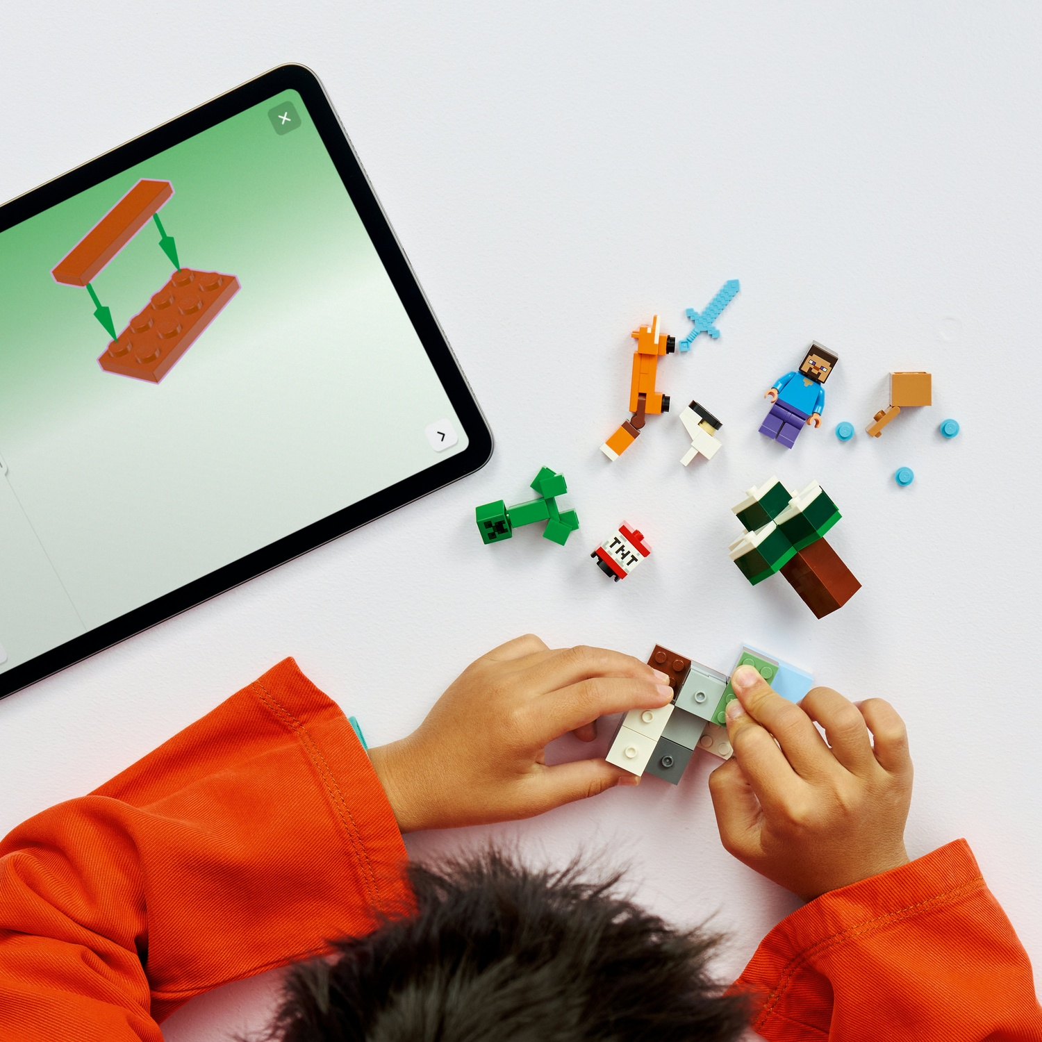Un bambino costruisce con i mattoncini Lego di Minecraft. Un tablet mostra le istruzioni. Varie figure e blocchi Lego su una superficie bianca.