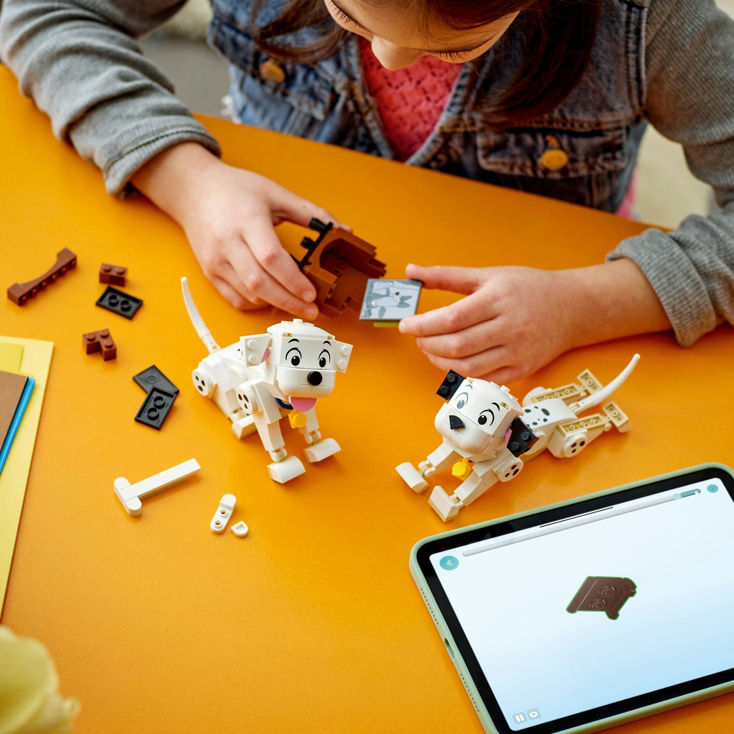 Un bambino sta costruendo figure Lego di dalmata su un tavolo arancione. Sono visibili due cani completati e pezzi Lego.