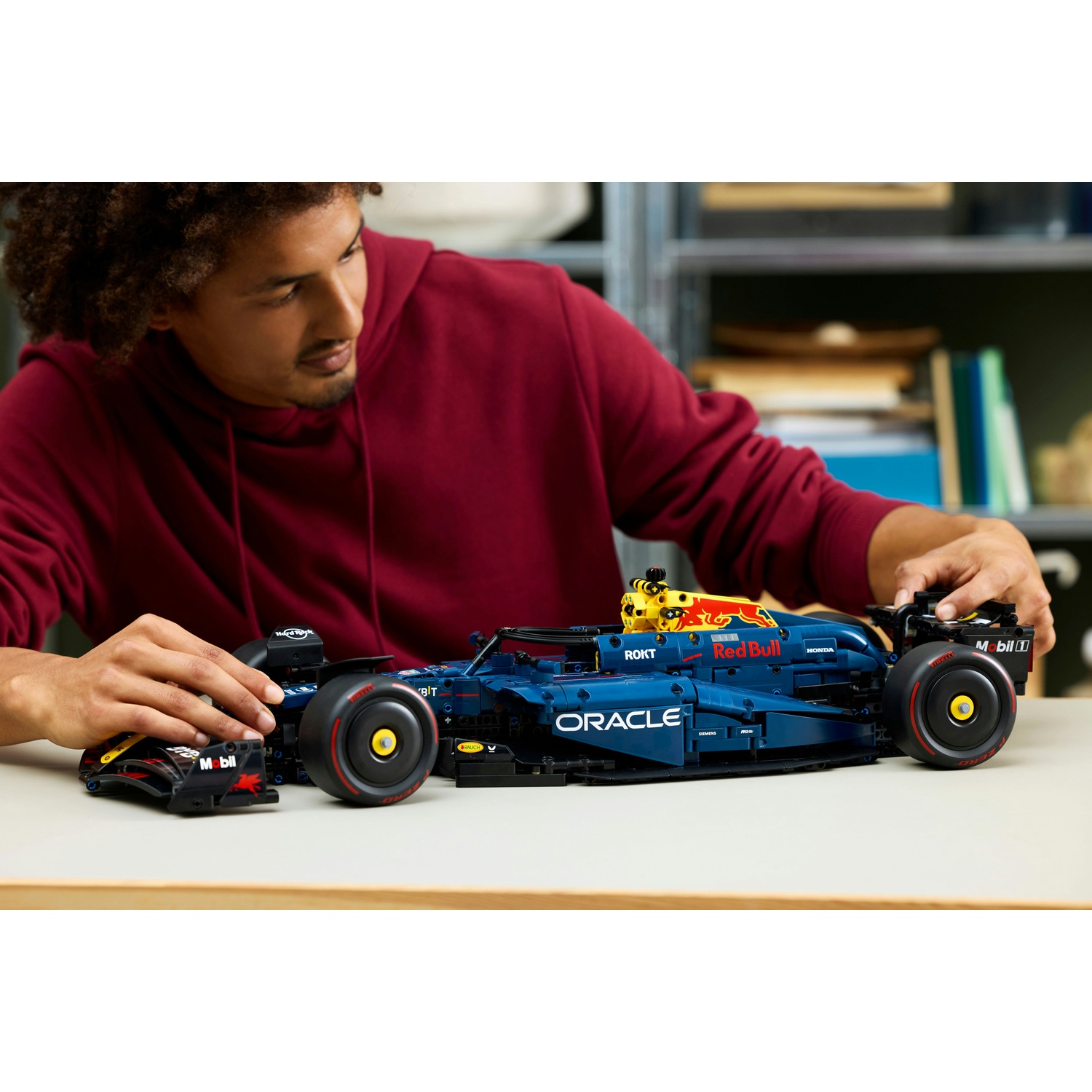 Un uomo con una felpa con cappuccio rossa assembla un'auto da corsa Lego Red Bull blu e gialla su un tavolo bianco.