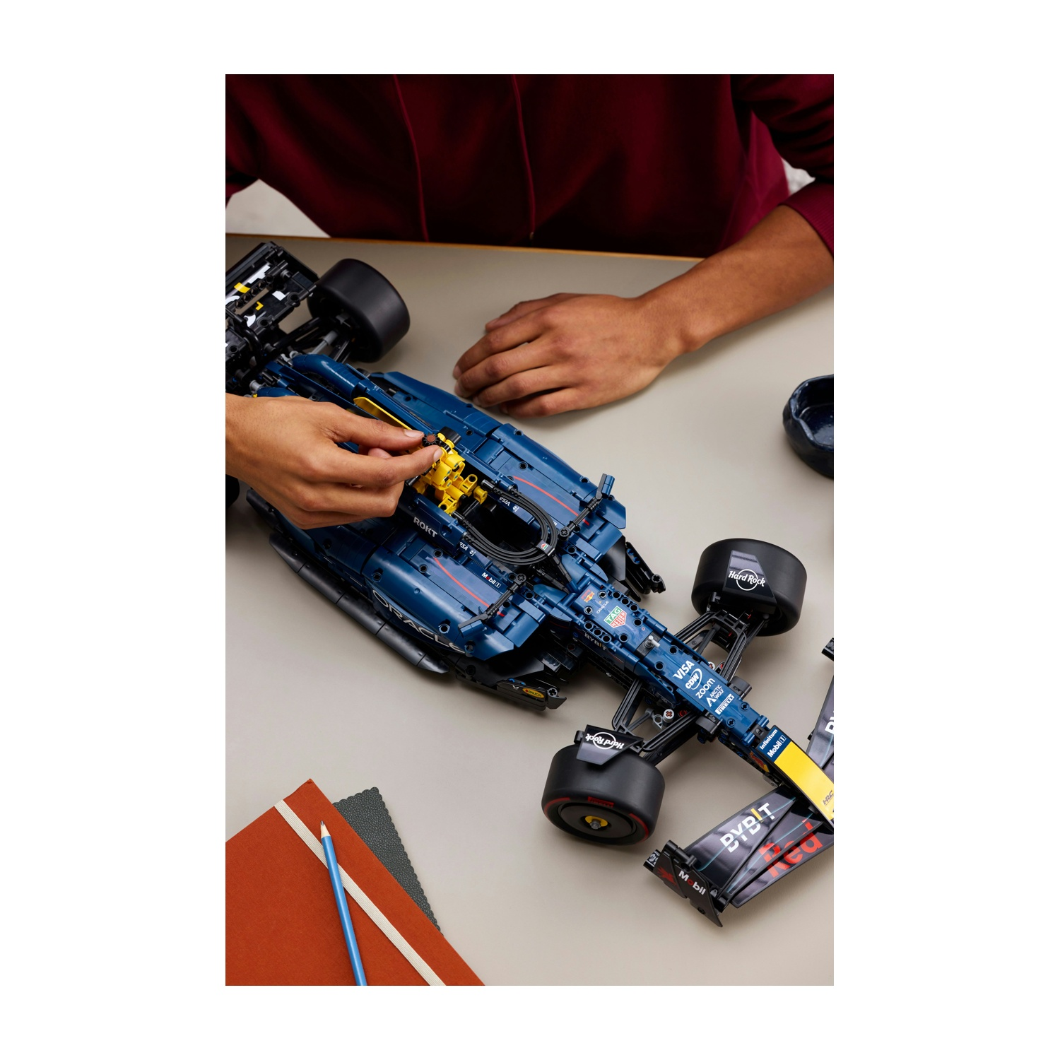 Mani che assemblano un modello di auto da corsa blu e gialla su un tavolo. Quaderni e una matita sono anche sul tavolo.