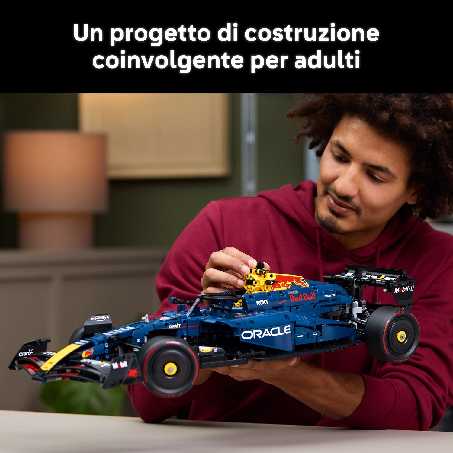 Un uomo assembla un'auto modello Red Bull F1 blu scuro. L'auto ha i loghi degli sponsor. L'uomo indossa una felpa con cappuccio bordeaux, al chiuso.