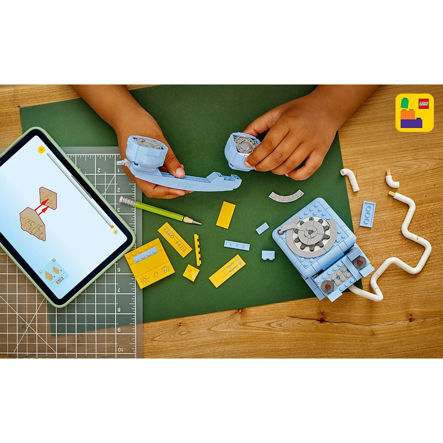 Mani che assemblano un set Lego blu su un tavolo di legno. Un tablet mostra le istruzioni. Pezzi Lego gialli e blu sparsi.